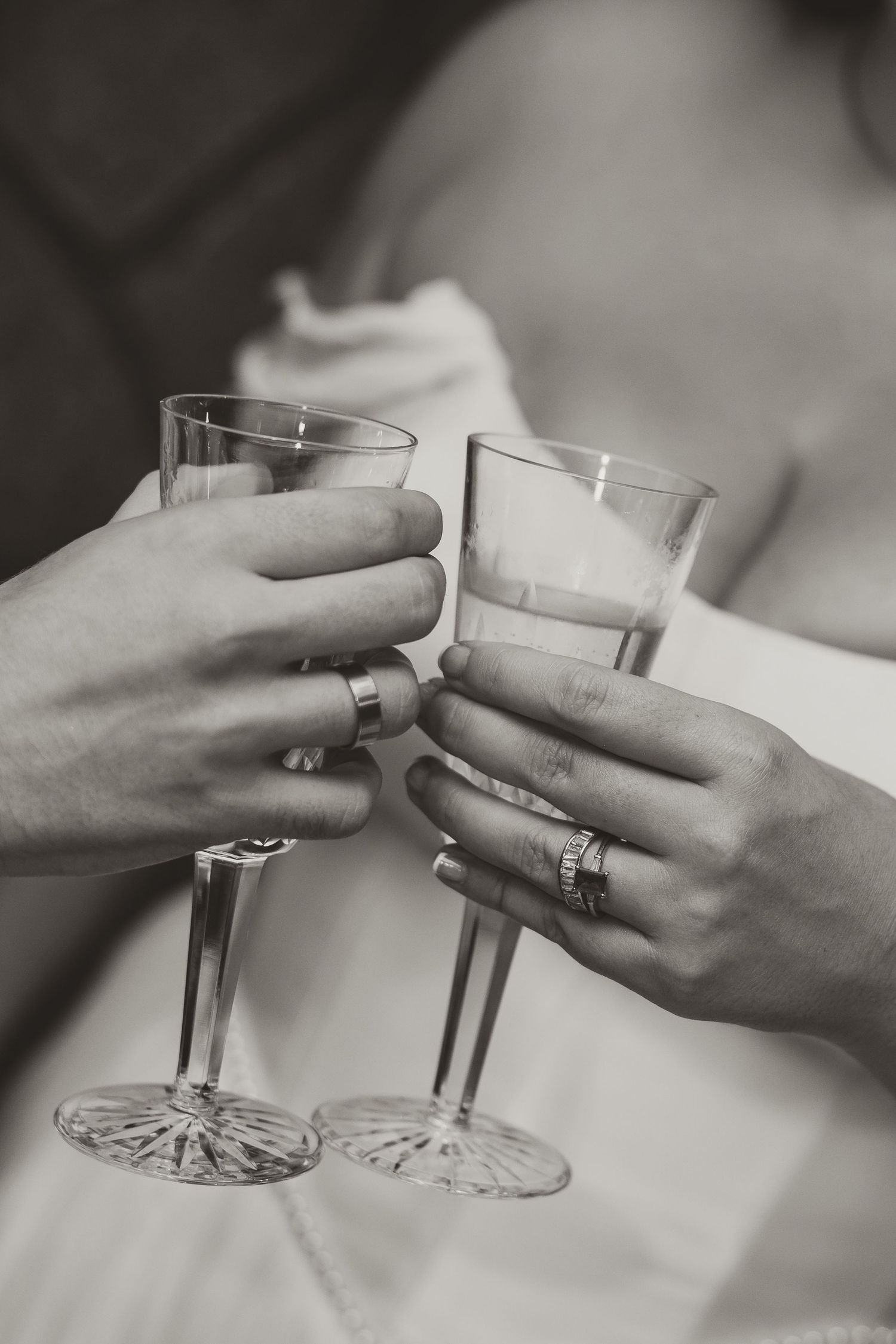 Black and white photo of two hands wearing rings clinking crystal champagne glasses in a wedding toast, with a bride in a white gown behind.
