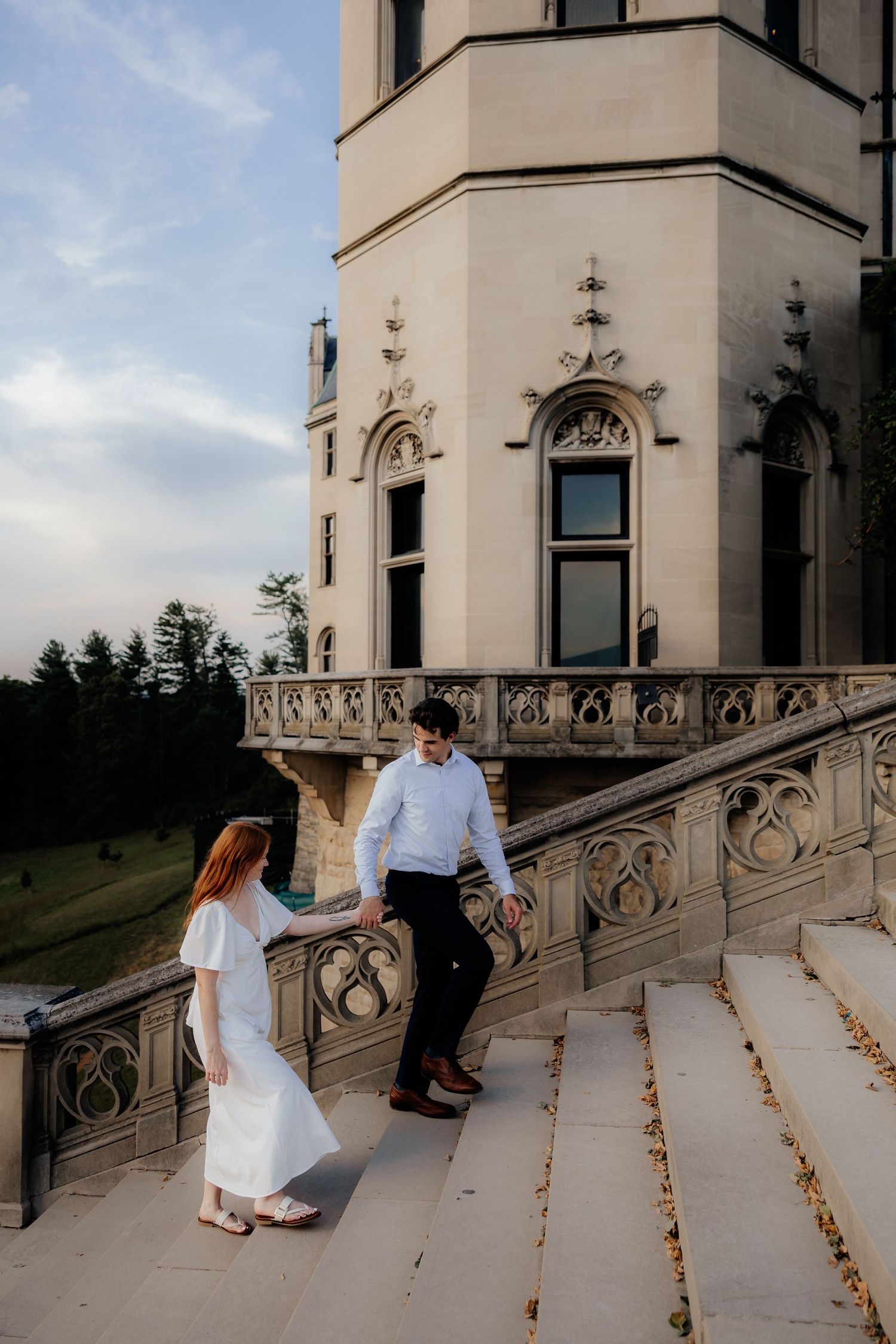 Couple in elegant attire holding hands on ornate castle steps at golden hour.