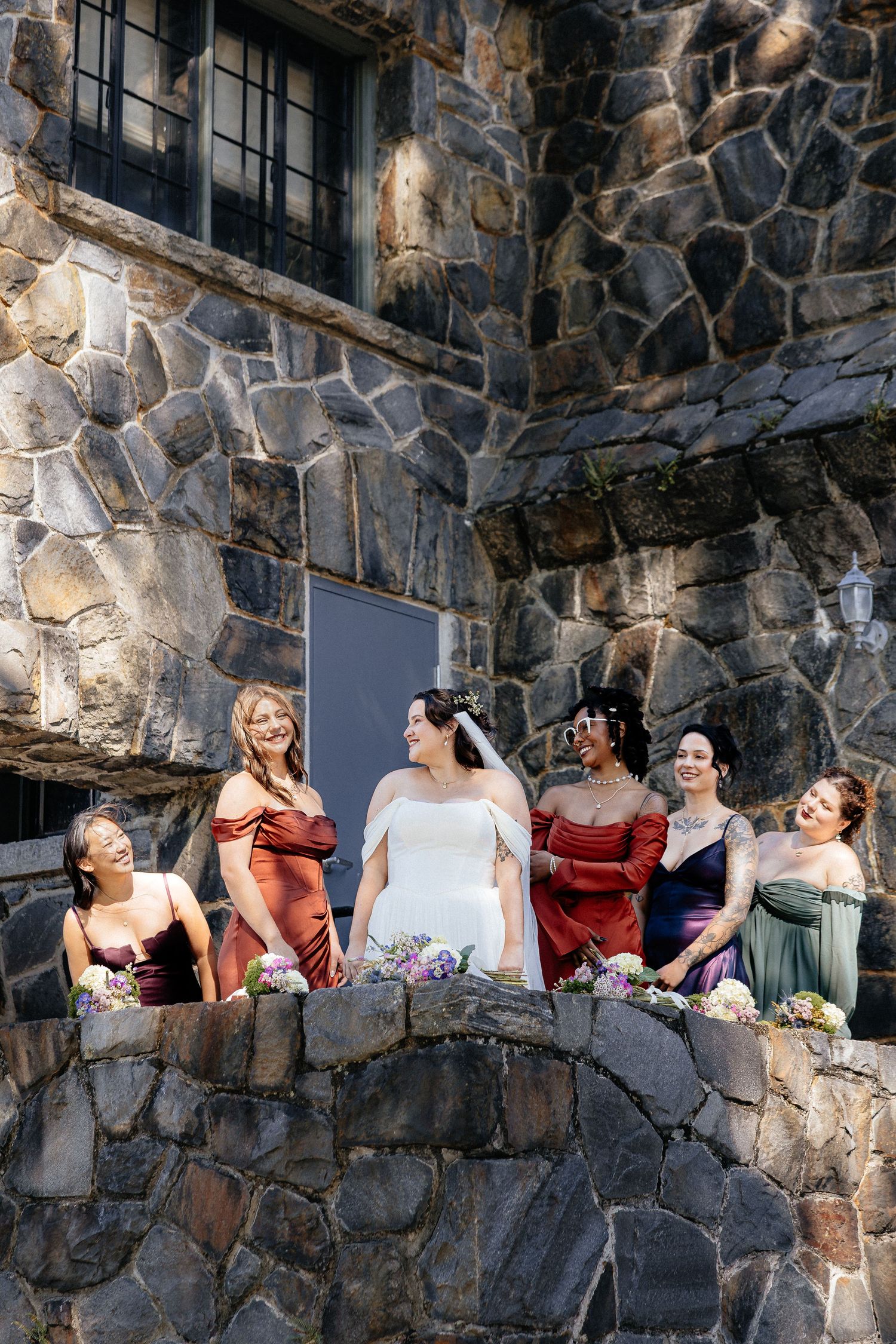 Bride in white gown stands with five bridesmaids in colorful mismatched dresses on a stone wall outside a rustic venue.