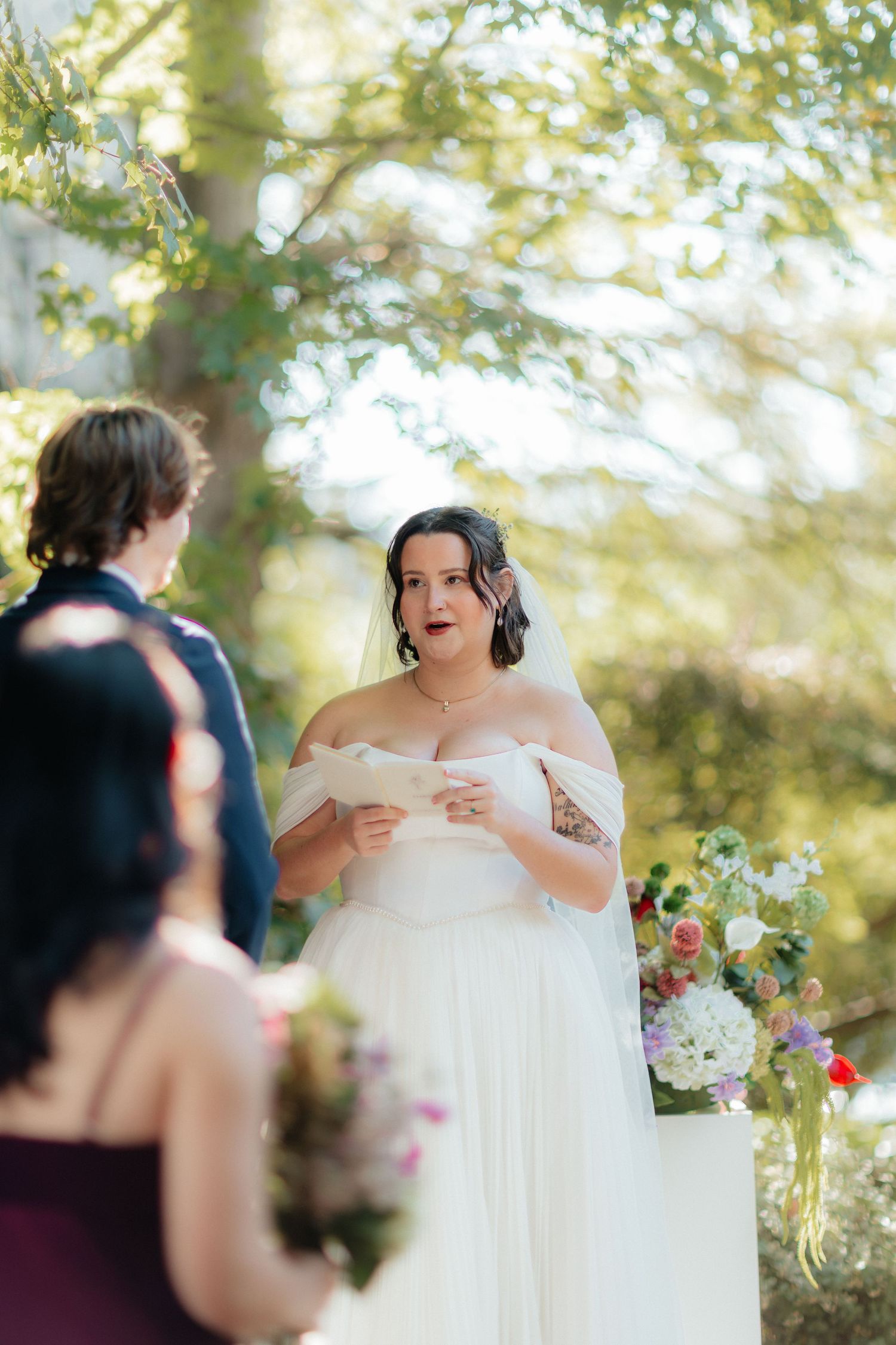 Bride in an off-the-shoulder white wedding gown reads her vows during an outdoor ceremony beside a colorful floral arrangement.