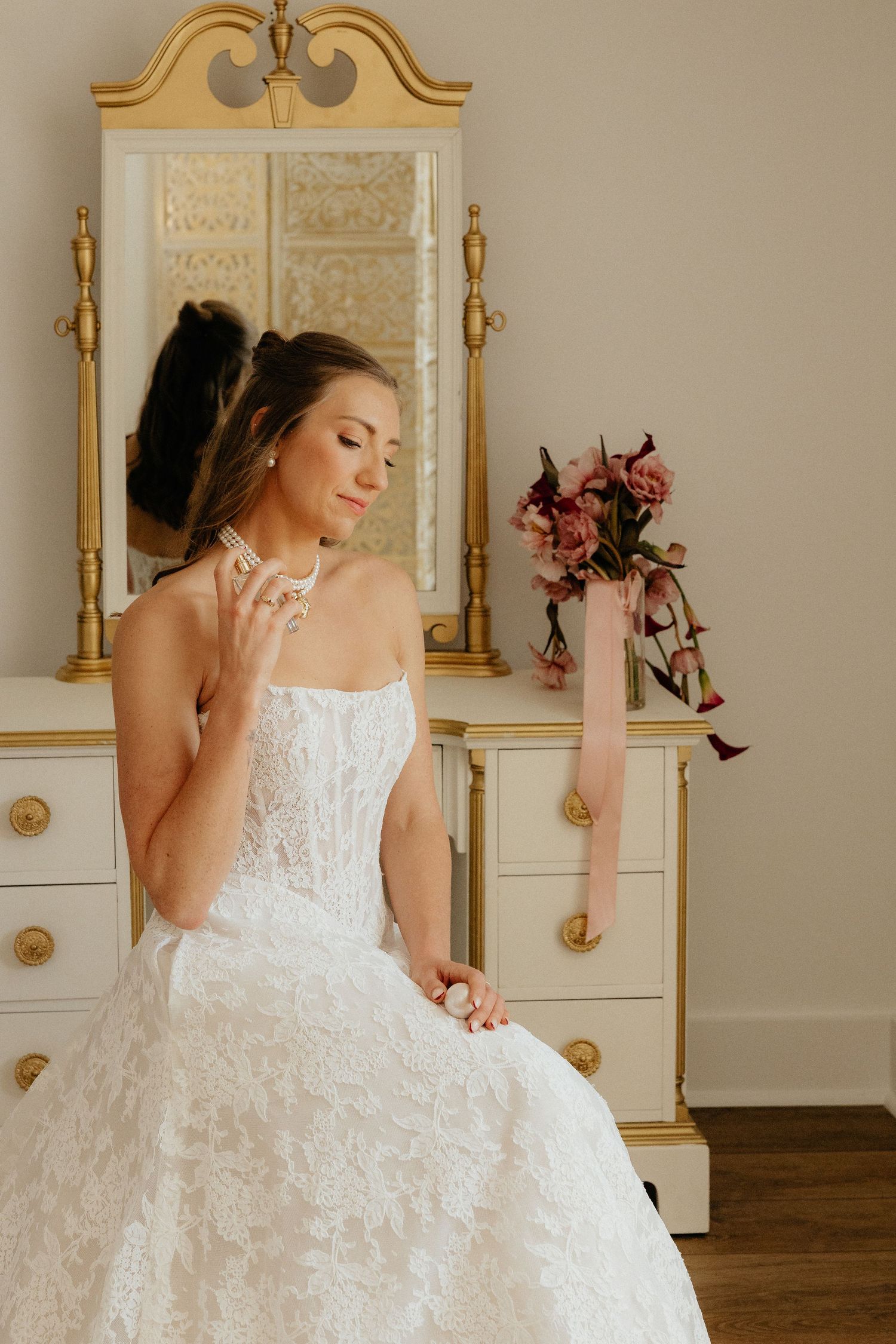 Bride in white lace gown sits by gold-framed mirror, holding necklace, with pink floral bouquet on dresser.