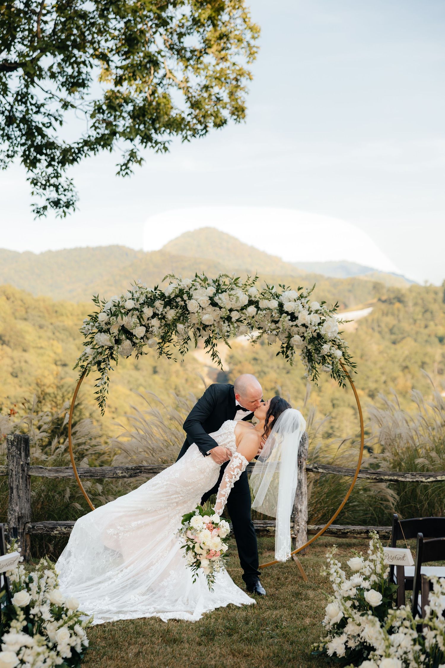Bride and groom share a dipped kiss beneath a floral wedding arch with mountain views and white bouquets in an outdoor ceremony.
