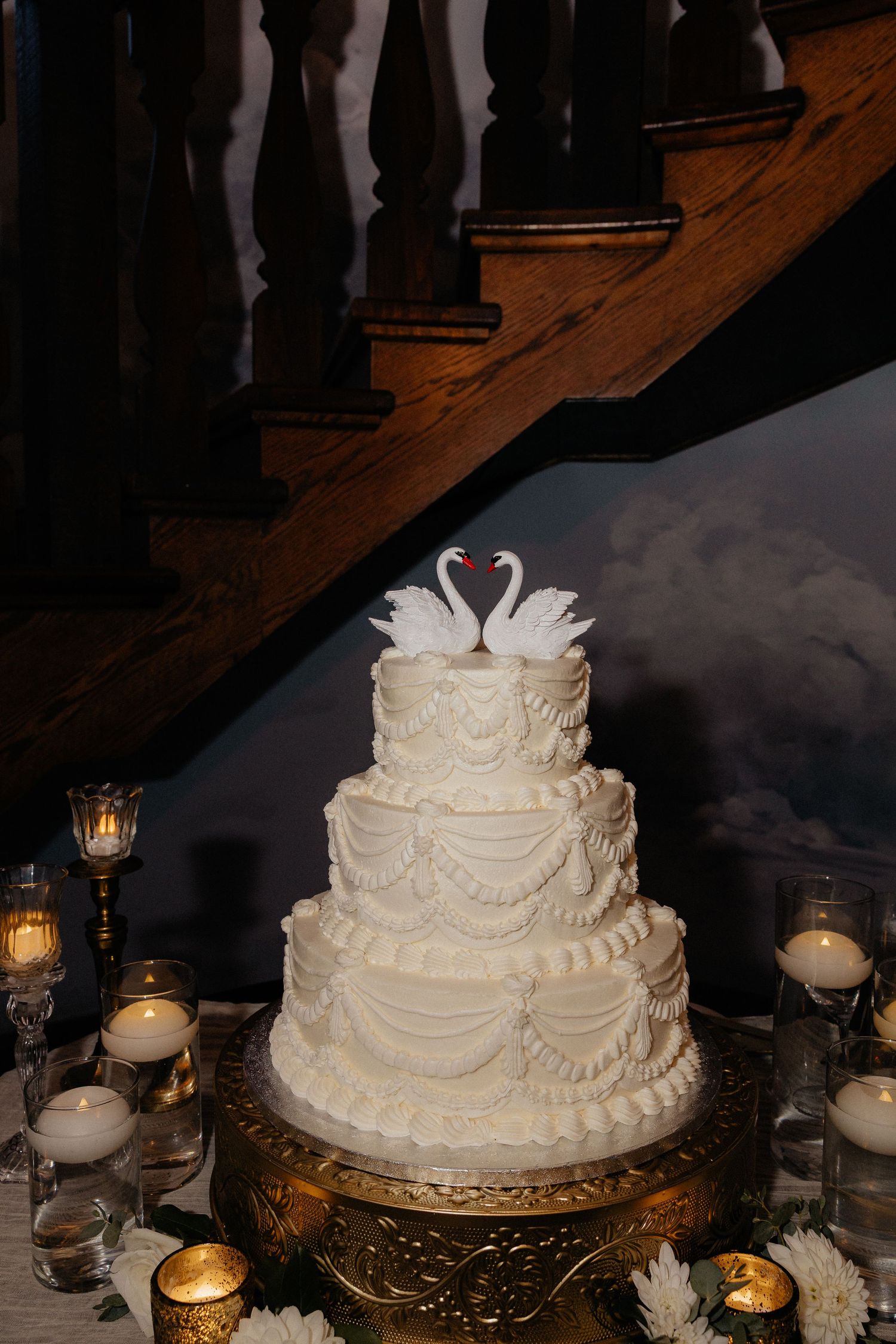 Three-tier white wedding cake with swan toppers on an ornate gold stand, surrounded by candles and flowers beneath a wooden staircase.