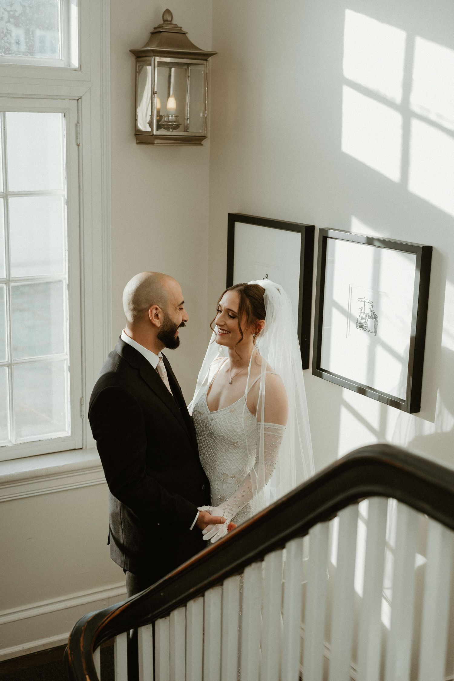 A couple shares an intimate moment on a bright staircase landing with a vintage lantern and natural light streaming in.
