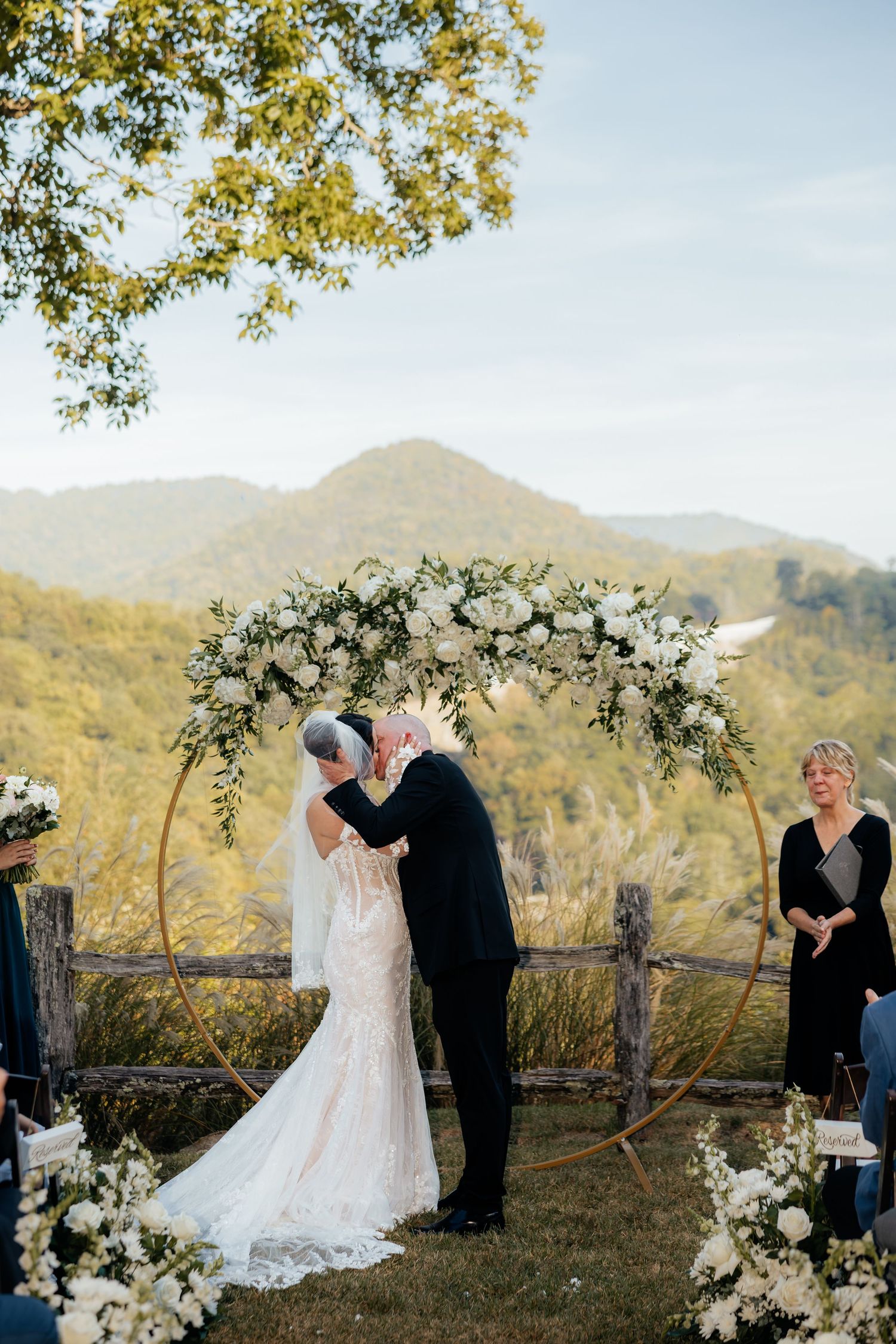 Bride and groom share a ceremony kiss beneath a round floral wedding arch with mountain views while an officiant looks on.