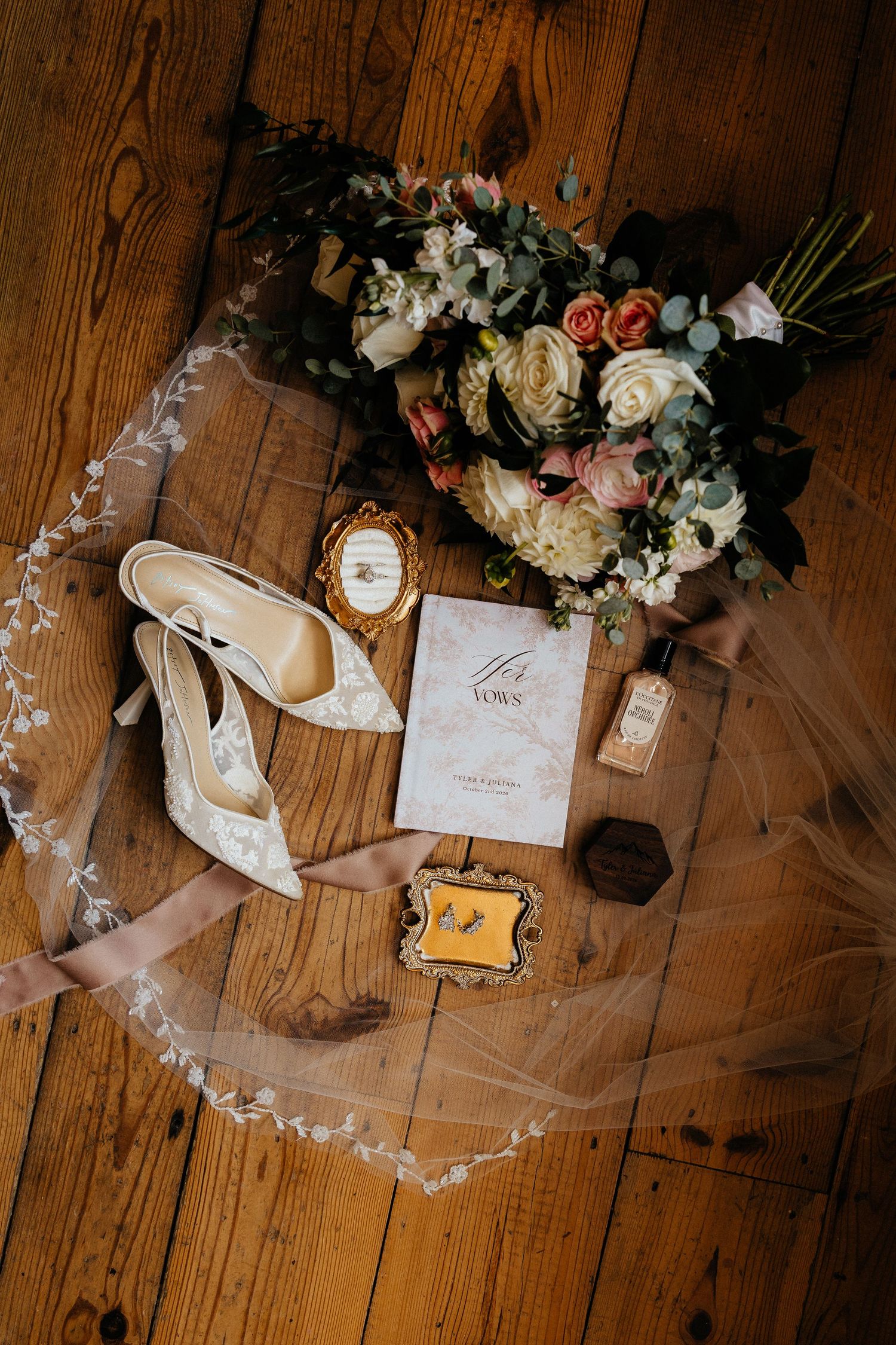 Wedding flat lay with a blush and ivory bouquet, lace bridal heels, a lace veil, vow booklet, and vintage frames on a rustic wooden floor.
