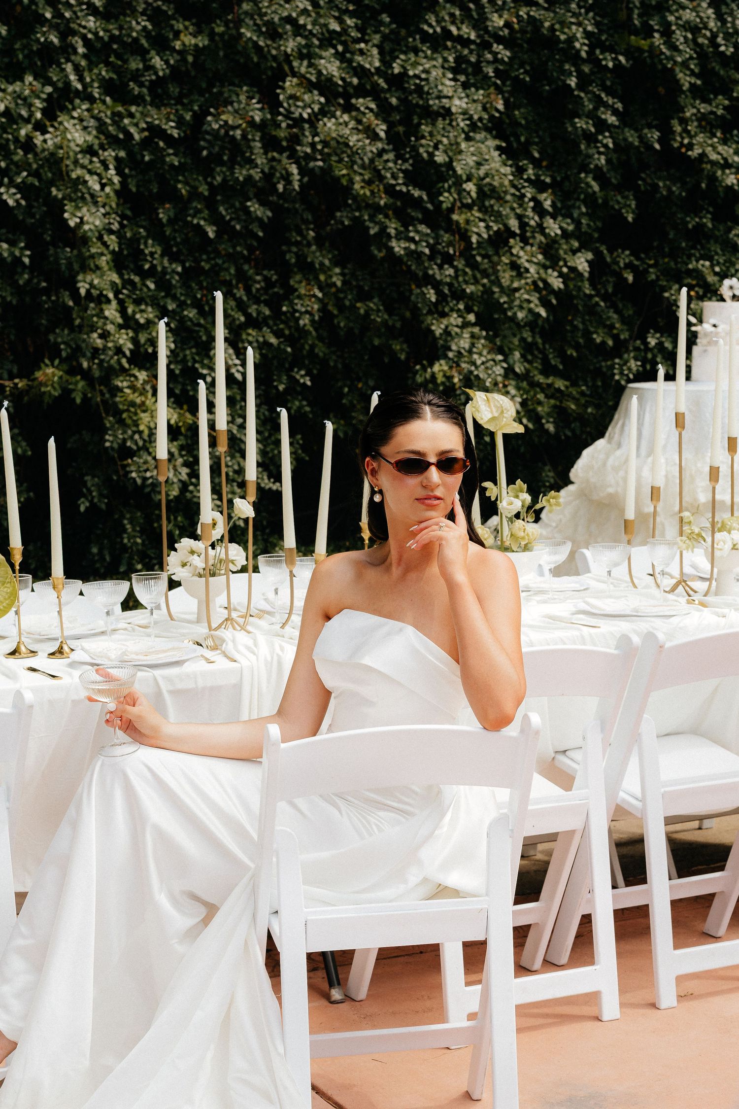 Bride in white strapless gown and sunglasses seated at elegant outdoor wedding reception table with gold candles.