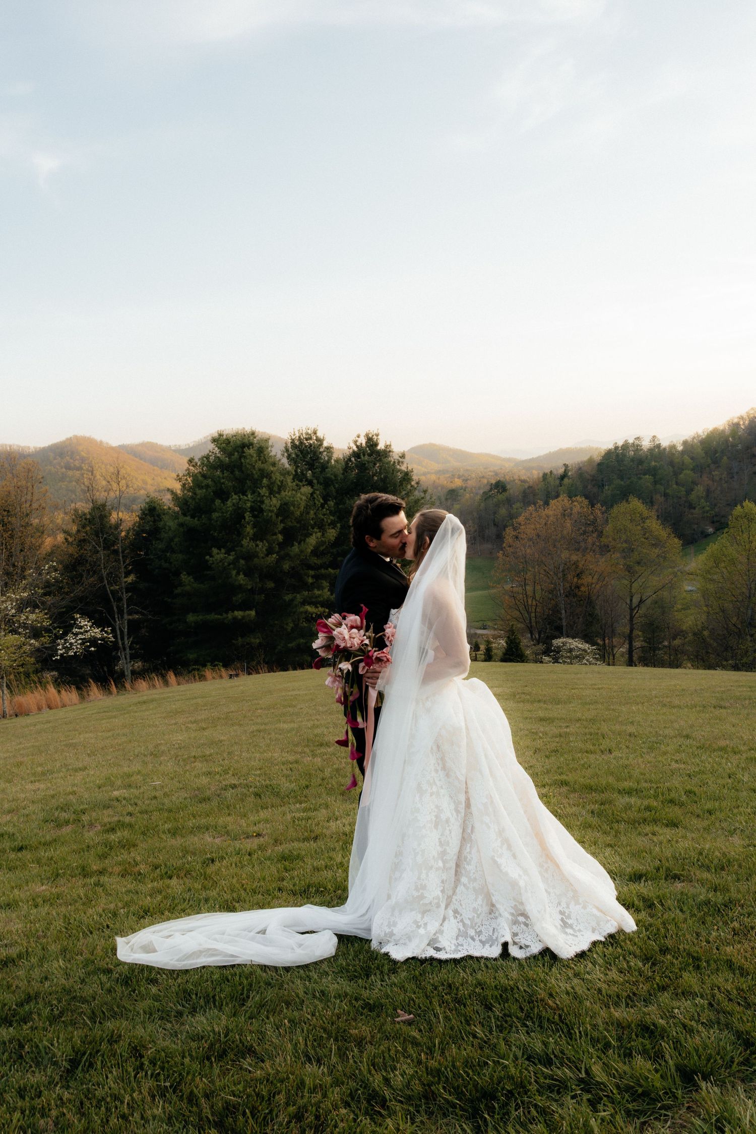 Bride in a lace wedding gown with a long veil kisses her groom on a grassy hilltop with mountains and autumn trees behind them.