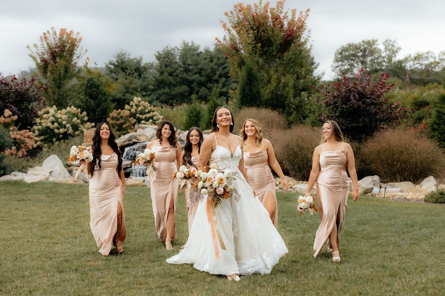 Bride in white gown walks with four bridesmaids in blush pink dresses on green lawn with lush garden backdrop.