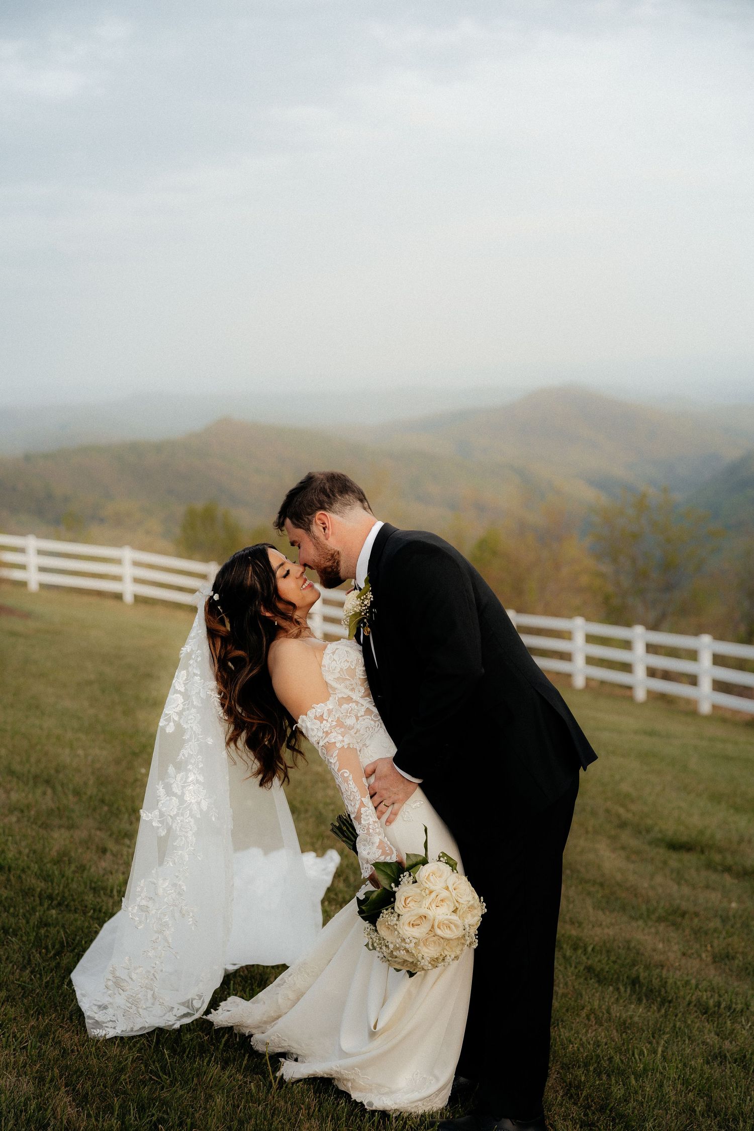 Bride and groom share a romantic kiss on a grassy hillside with stunning mountain views and white fence backdrop.