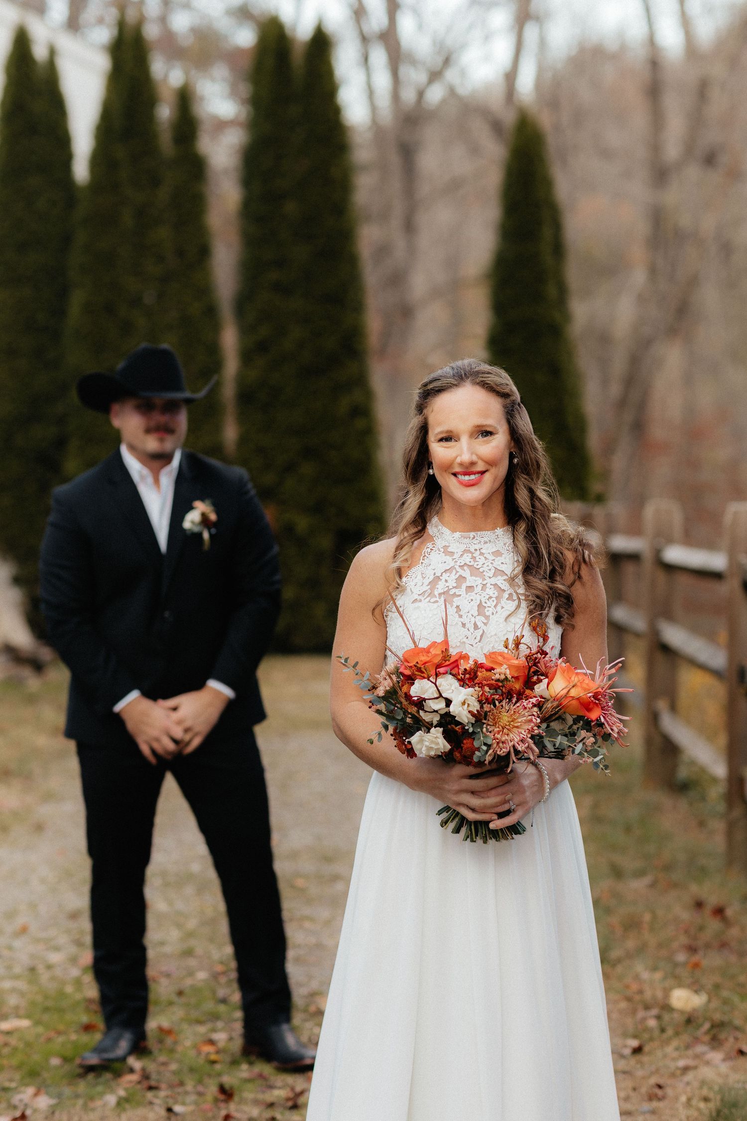 Smiling bride in white lace gown holds orange bouquet outdoors, groom in black suit and cowboy hat stands behind her.