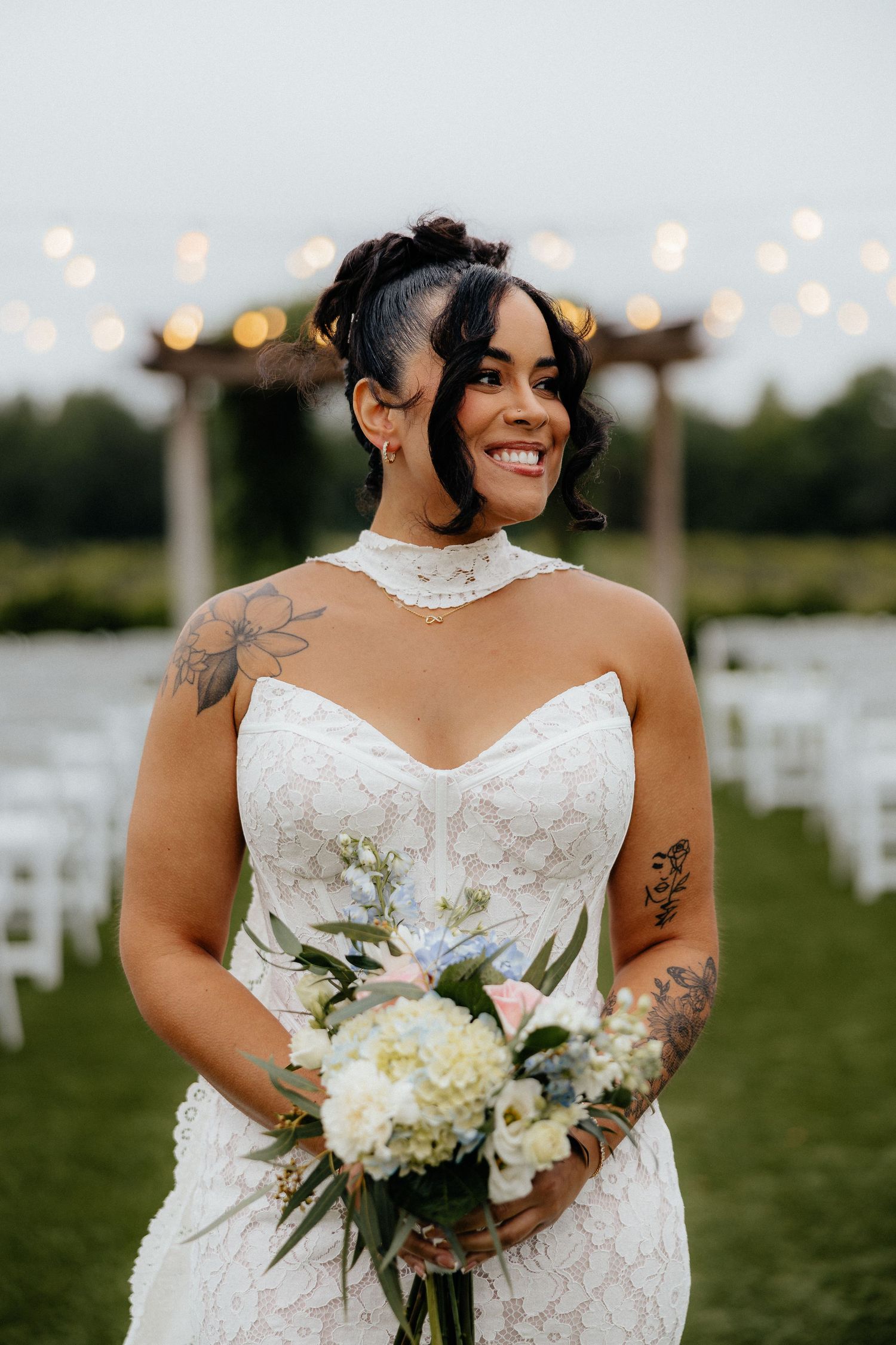 Smiling bride in white lace halter gown holding pastel bouquet, tattoos visible, outdoor wedding ceremony with string lights.