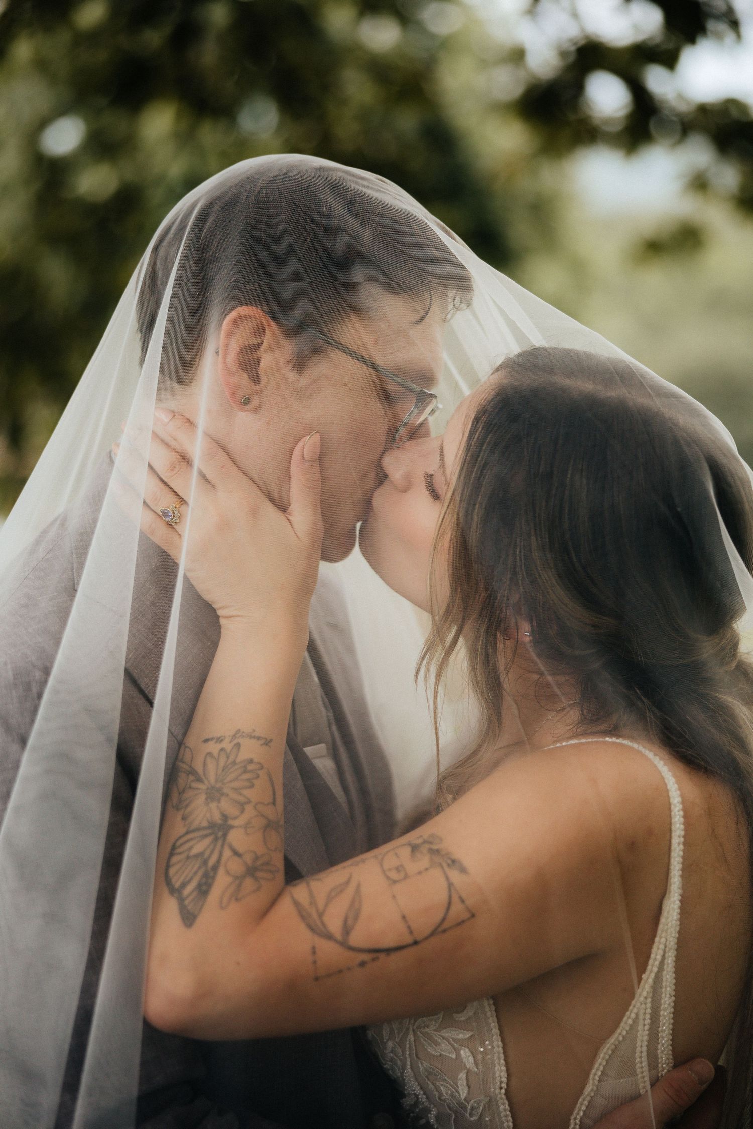 A bride and groom share a romantic kiss under a wedding veil outdoors, the bride showcasing floral tattoos on her arms.
