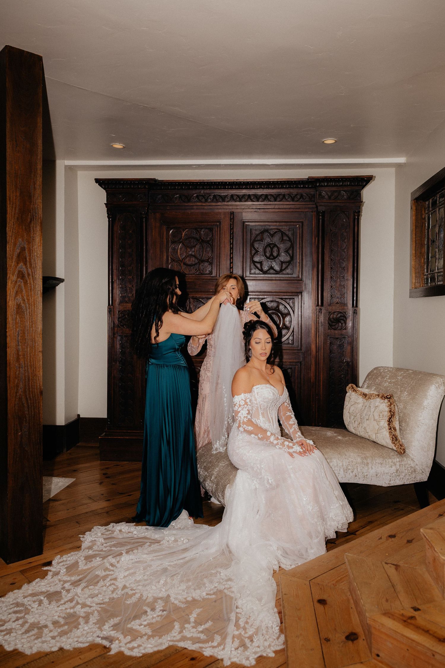 Bride in a lace long-sleeve wedding gown with a long train sits on a chaise while a bridesmaid in teal helps secure her veil.