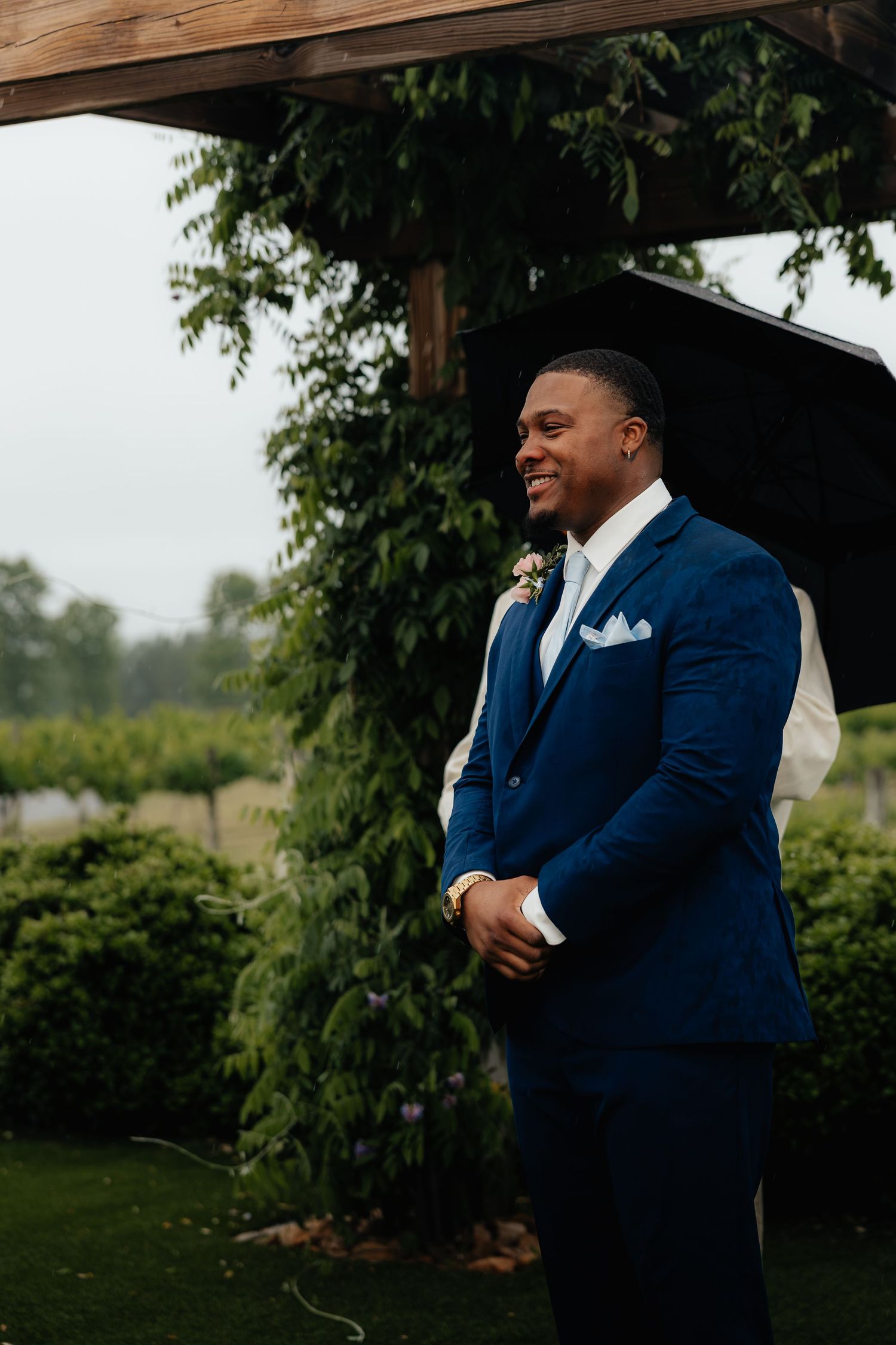Smiling groom in navy blue suit with boutonniere standing outdoors under pergola on rainy wedding day.