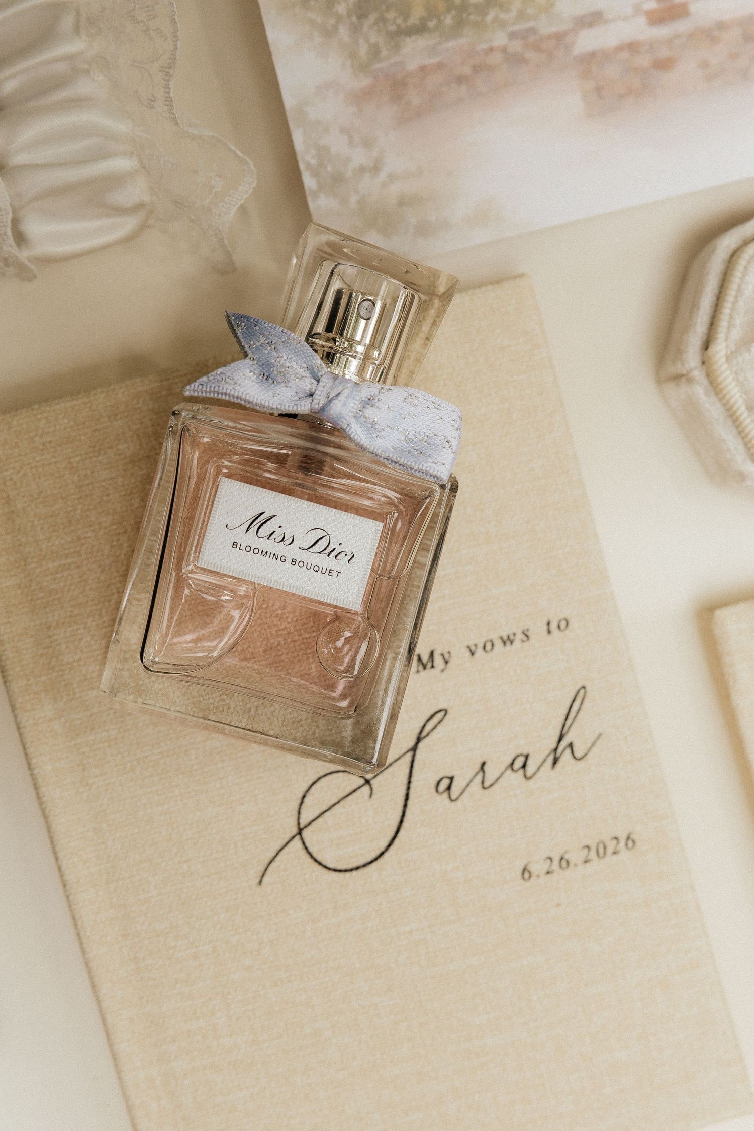 Miss Dior Blooming Bouquet perfume with a blue ribbon rests on a cream wedding vow book labeled My vows to Sarah, dated 6.26.2026.