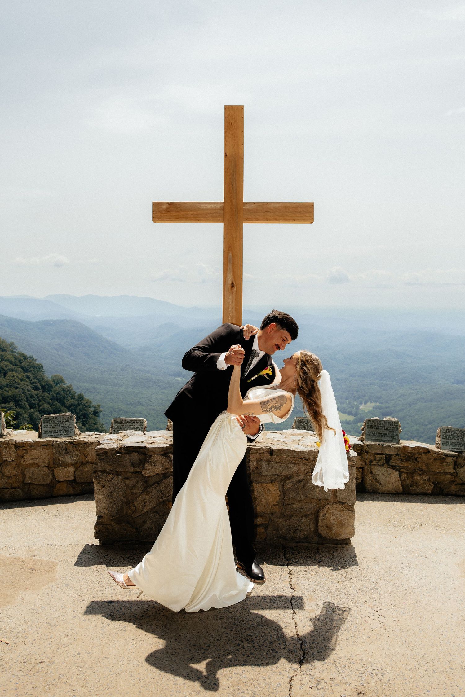 Bride and groom share a dramatic dip kiss beside a stone wall and large wooden cross with a scenic mountain view behind them.