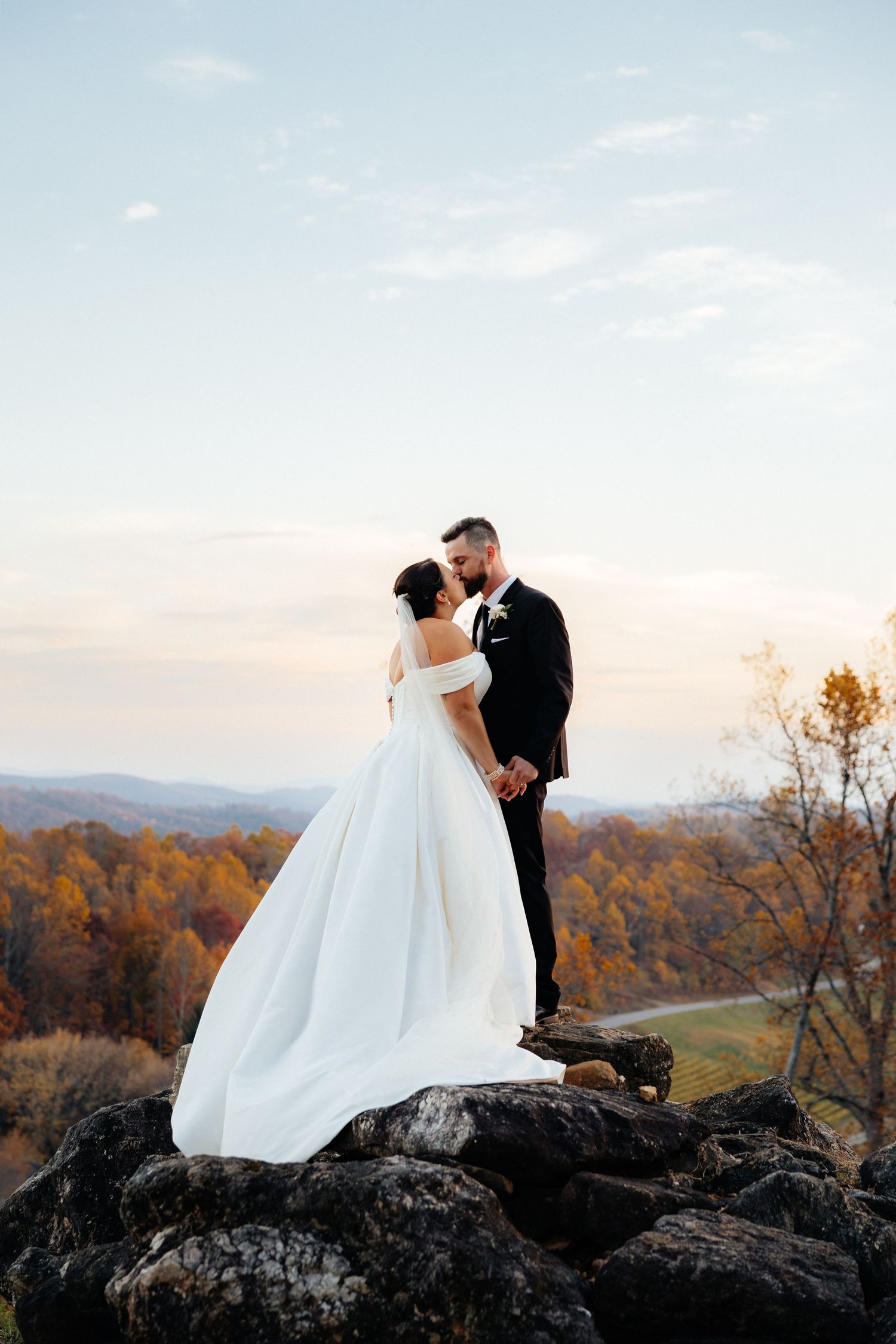 Newlywed couple embraces on rocky mountain overlook during autumn sunset with colorful fall foliage in background.