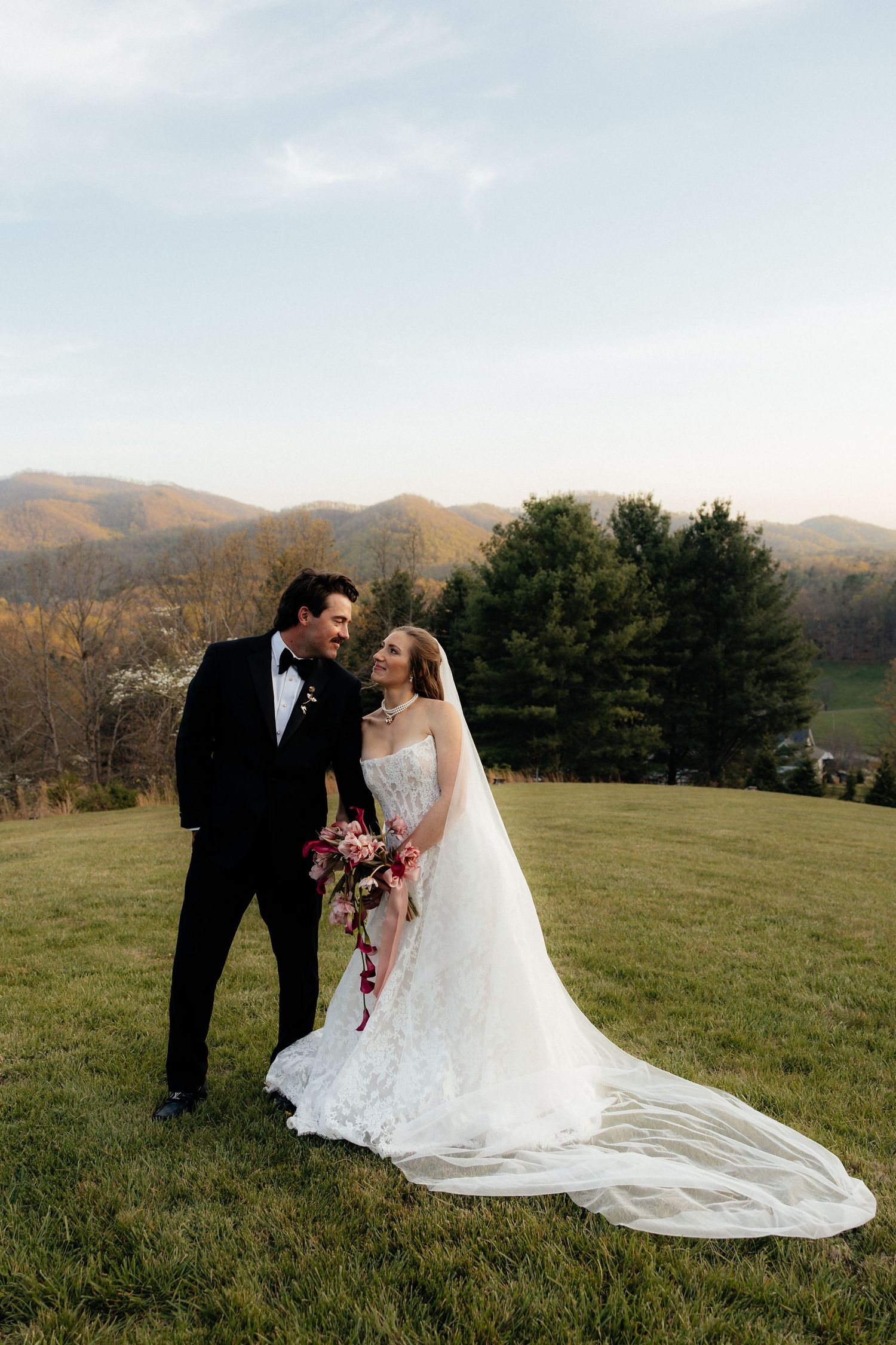 Bride in a lace gown with a long veil and groom in a black tuxedo hold a bouquet on a grassy hill with mountains behind.