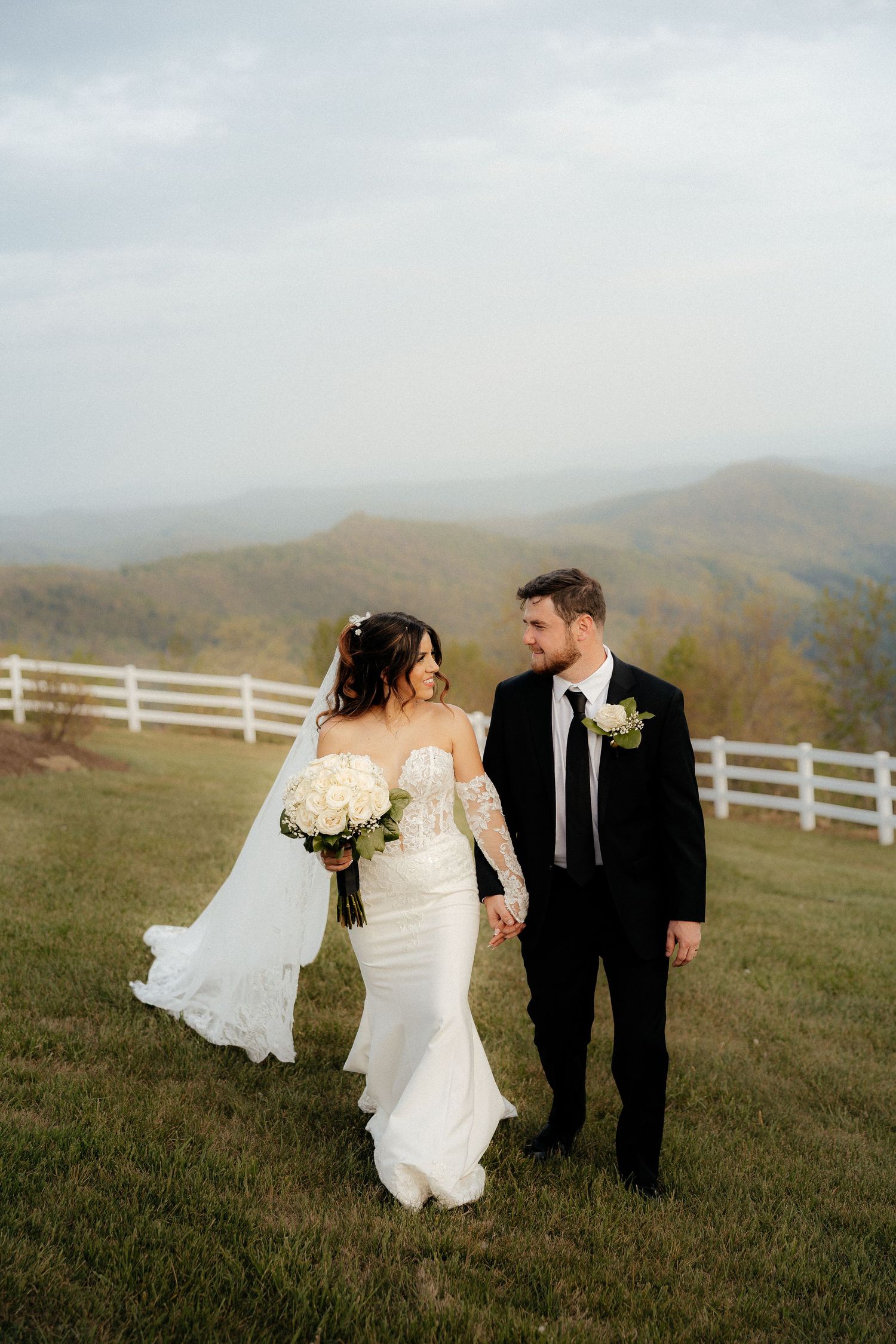 Bride and groom holding hands walking on a grassy hilltop with mountain views and white fence backdrop.