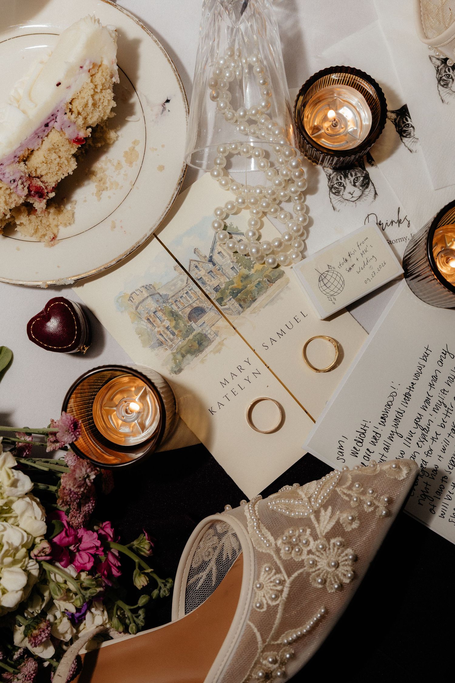 Flat lay of wedding details with a cake slice, pearl necklace, gold rings, illustrated invitation, lit candles, flowers and a beaded heel.