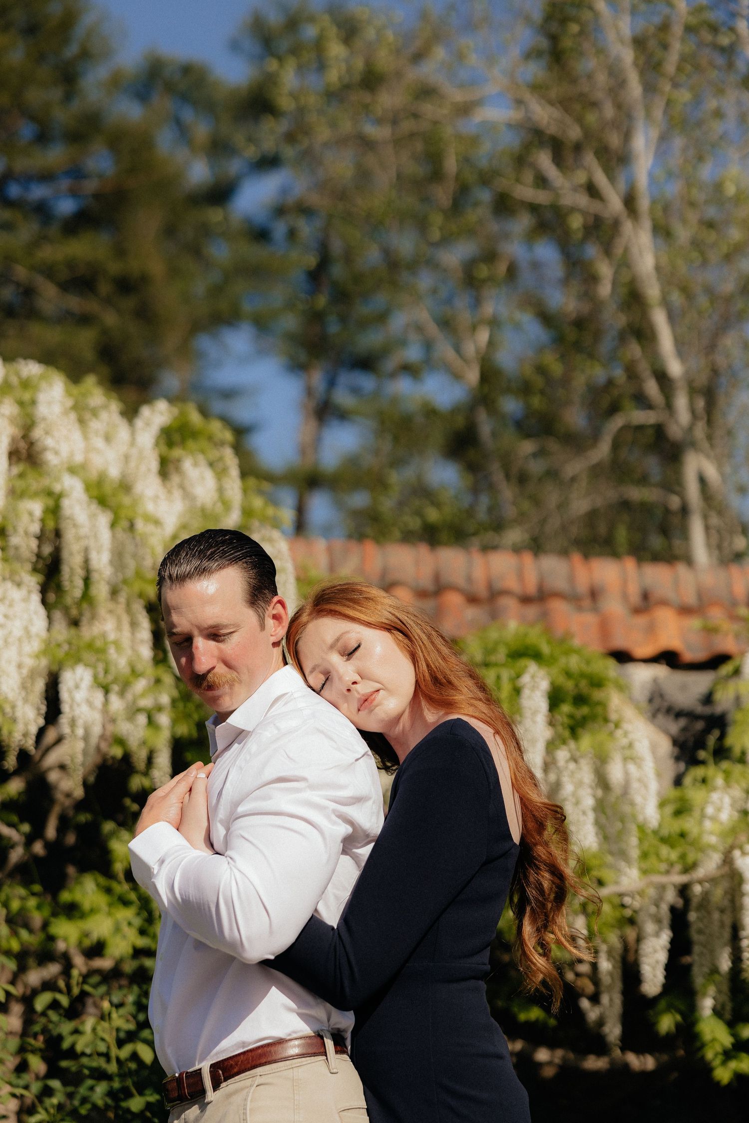 A couple embraces outdoors in a garden setting with white wisteria flowers and trees in the background.