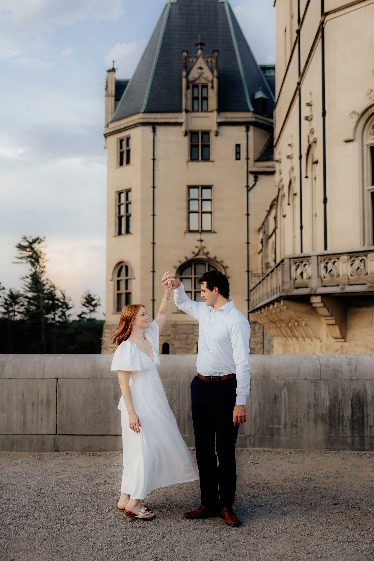 Couple dancing elegantly in front of a grand château, woman in white dress, man in white shirt and black pants.
