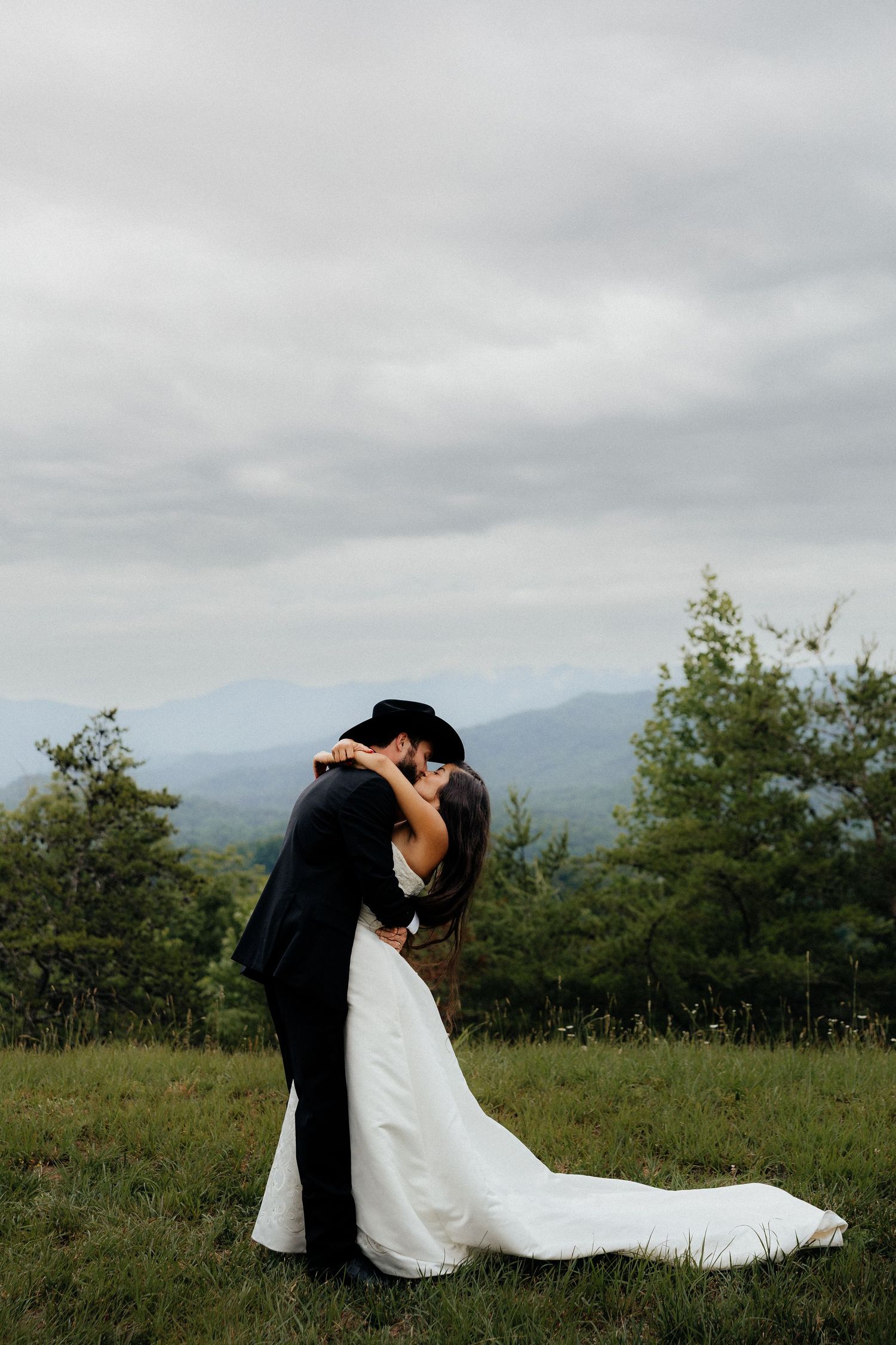 A bride and groom share a romantic kiss in a lush green mountain landscape under a cloudy sky.