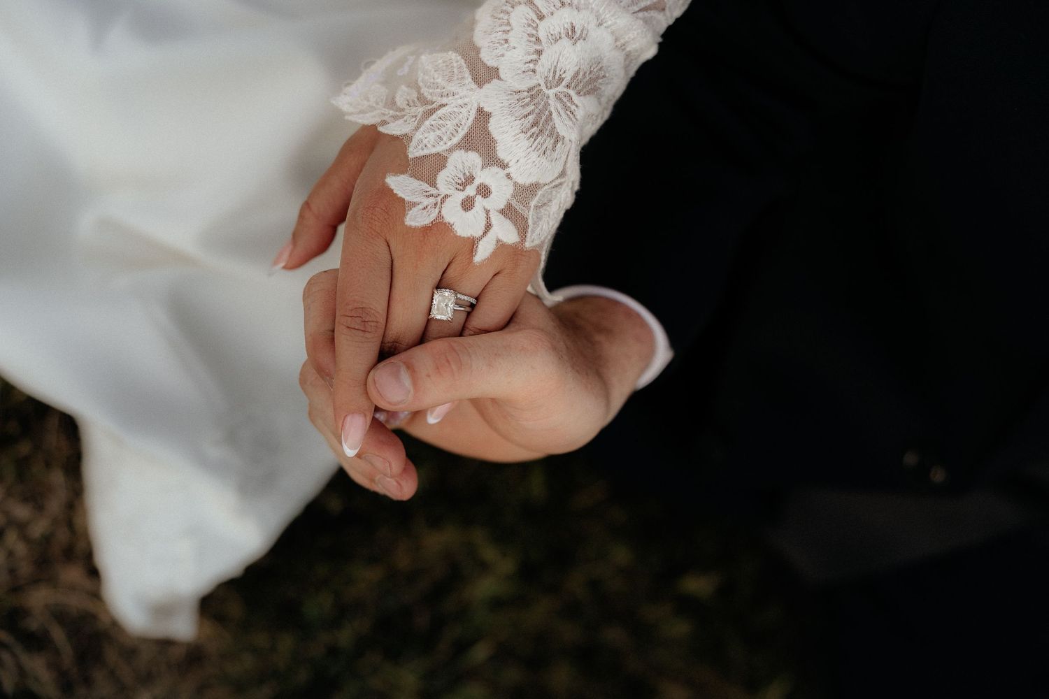 Bride and groom holding hands, showcasing elegant lace wedding dress sleeve and diamond engagement ring.