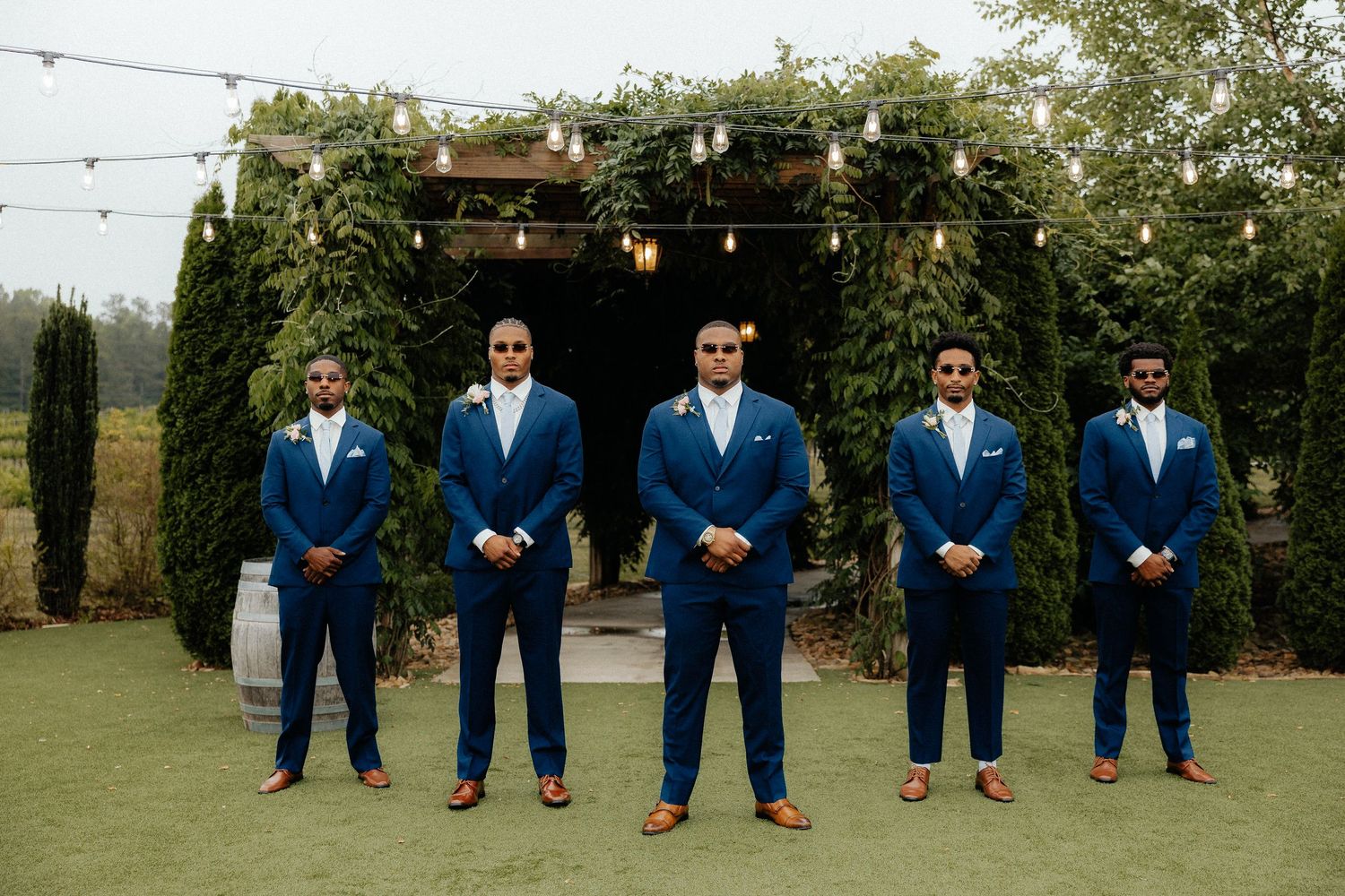Five groomsmen in matching navy blue suits with brown shoes stand outdoors at a wedding venue with string lights.