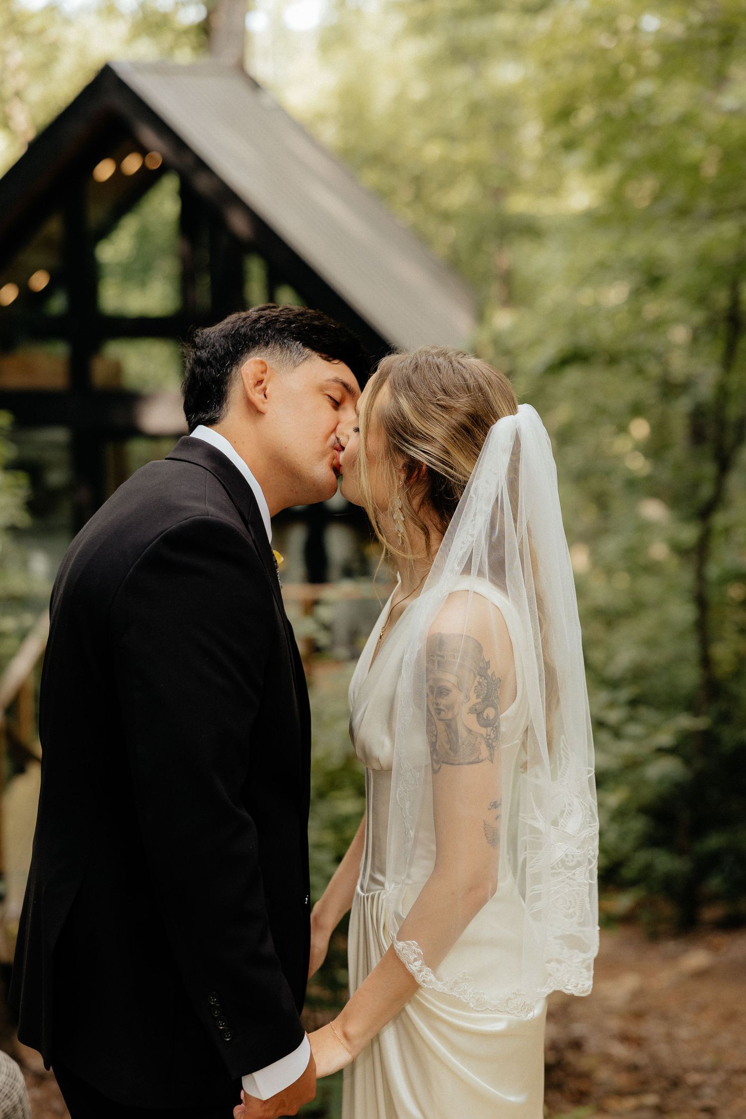 Bride in a lace veil and groom in a dark suit share a kiss during their woodland wedding ceremony near a rustic wooden chapel.