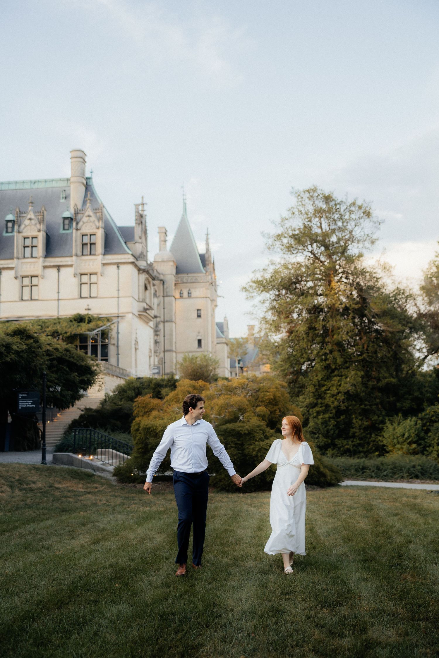 Couple holding hands walking on green lawn in front of a grand castle-like estate at dusk.