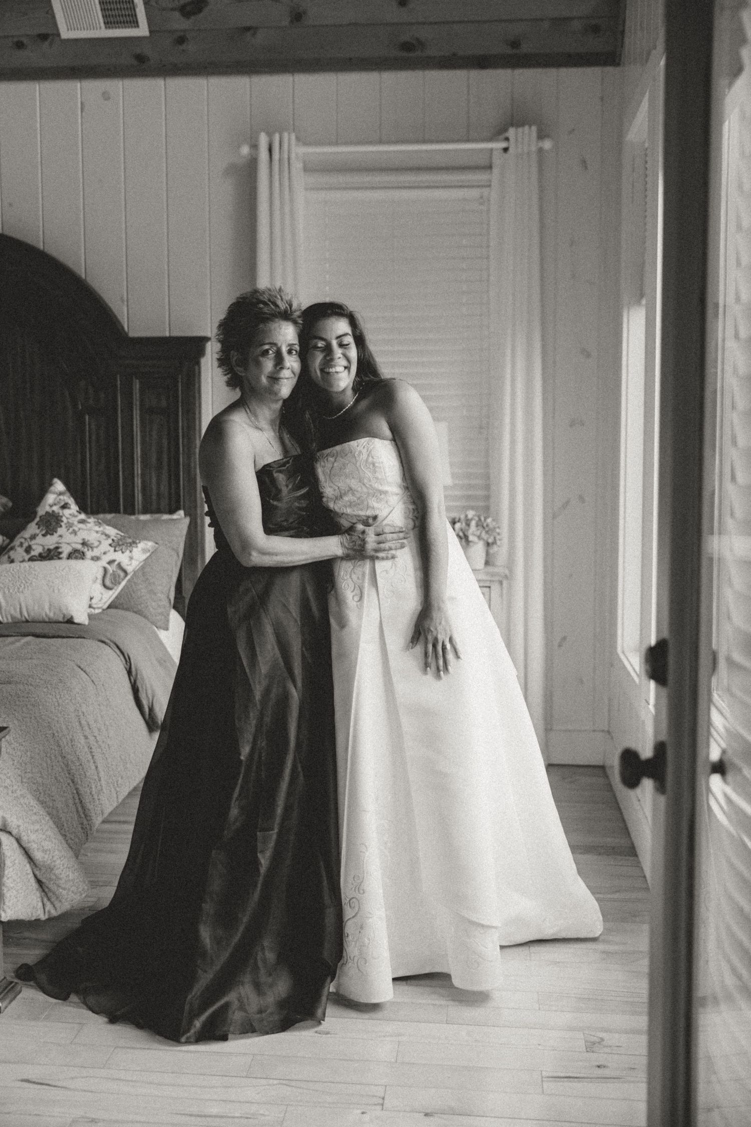 Black and white photo of bride in white gown embracing bridesmaid in dark dress inside rustic bedroom.