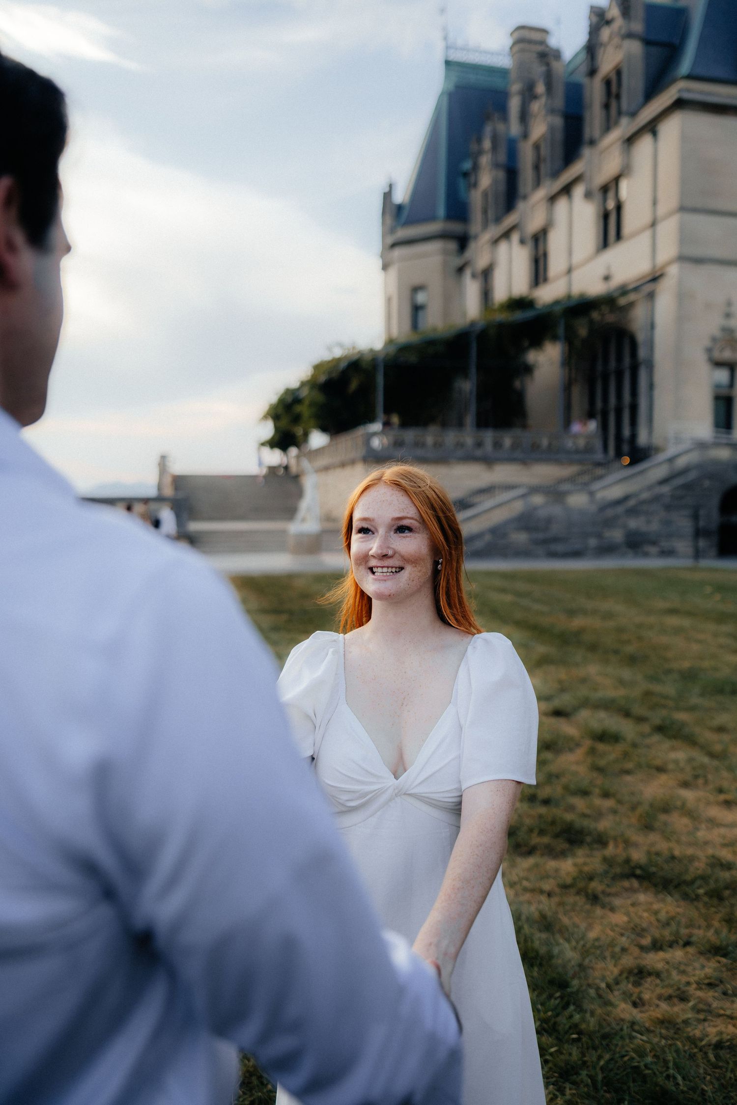 Smiling red-haired bride in white gown holds groom's hand before historic Biltmore Estate.