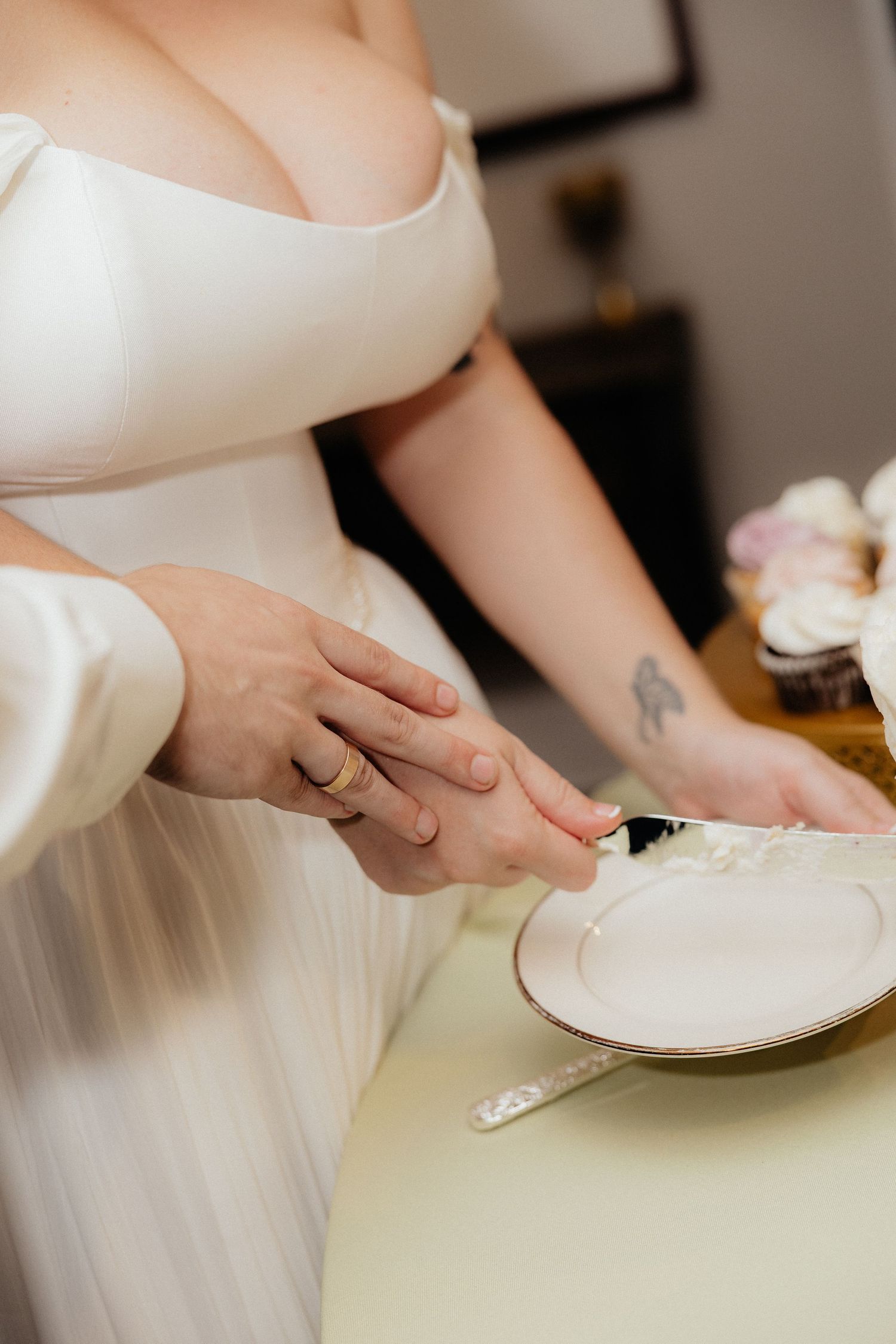 Bride in a white off-the-shoulder gown and a partner's hands cutting the wedding cake together with a knife over a plate.