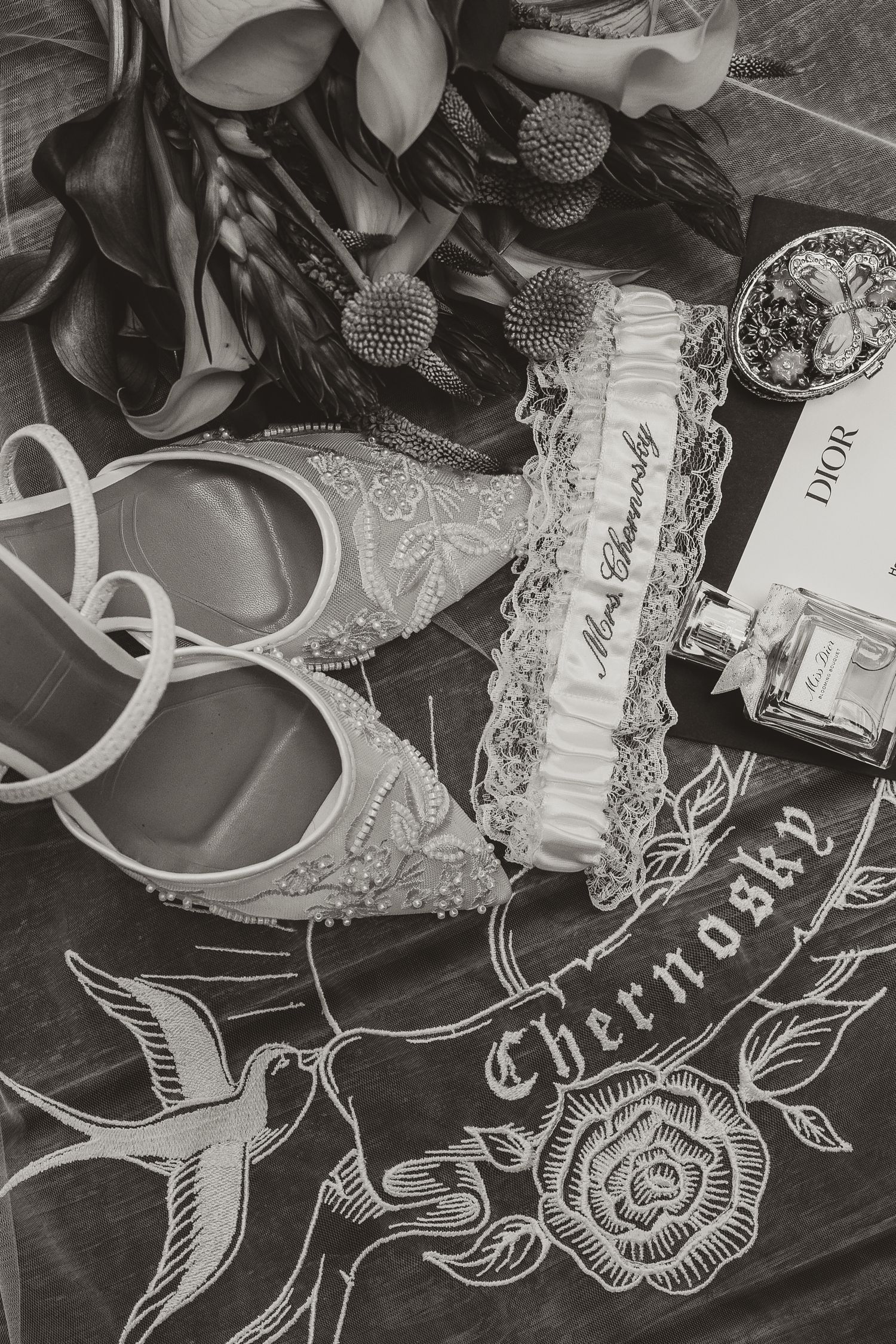 Black and white wedding flat lay with beaded bridal shoes, a lace garter, Dior perfume, a floral bouquet and a swallow rose design.