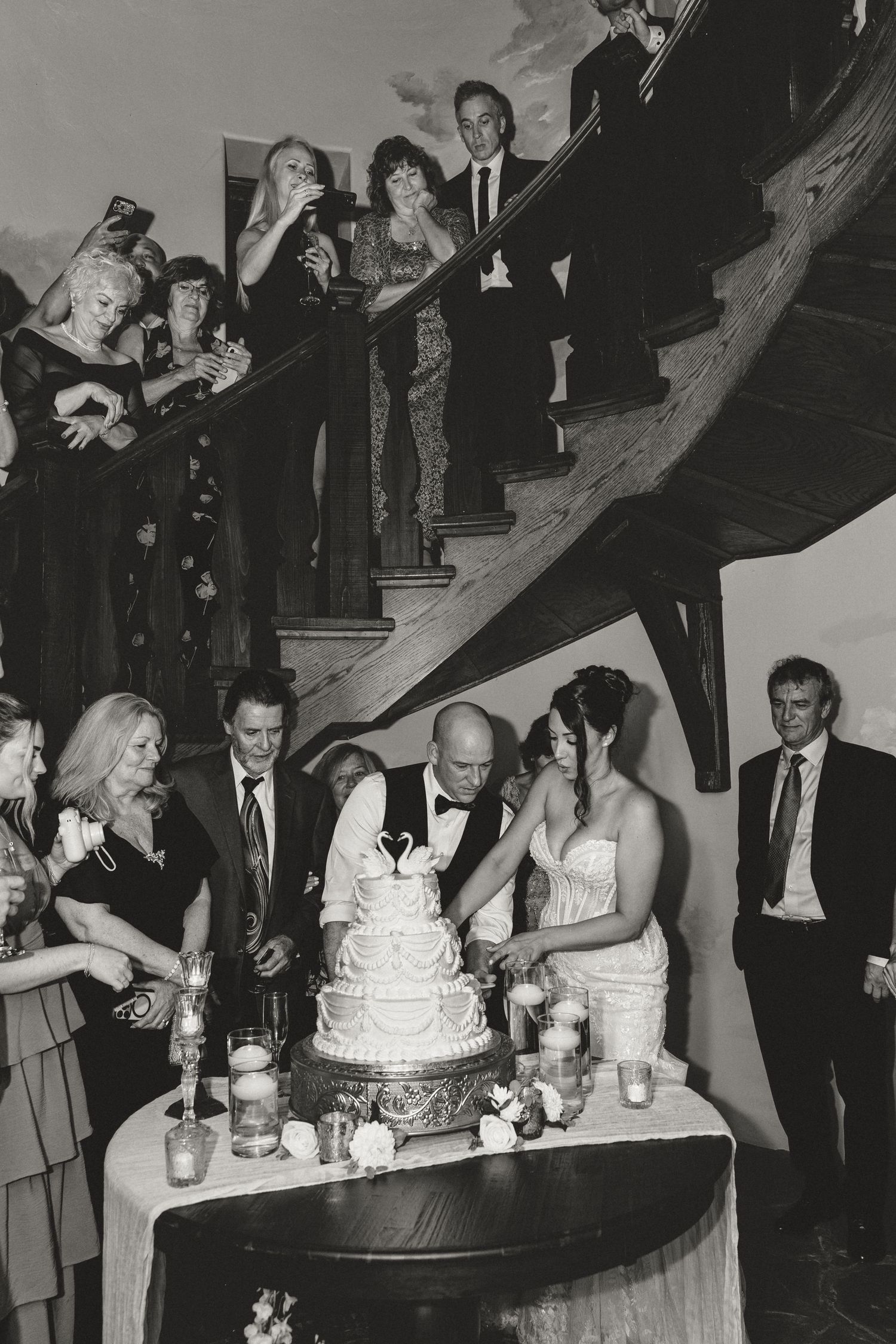 Black and white wedding photo of a bride and groom cutting a tiered cake with swan topper while guests watch from a grand staircase.