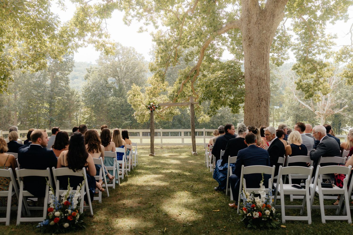 Outdoor wedding ceremony with guests seated in white chairs on a lawn surrounded by tall trees.