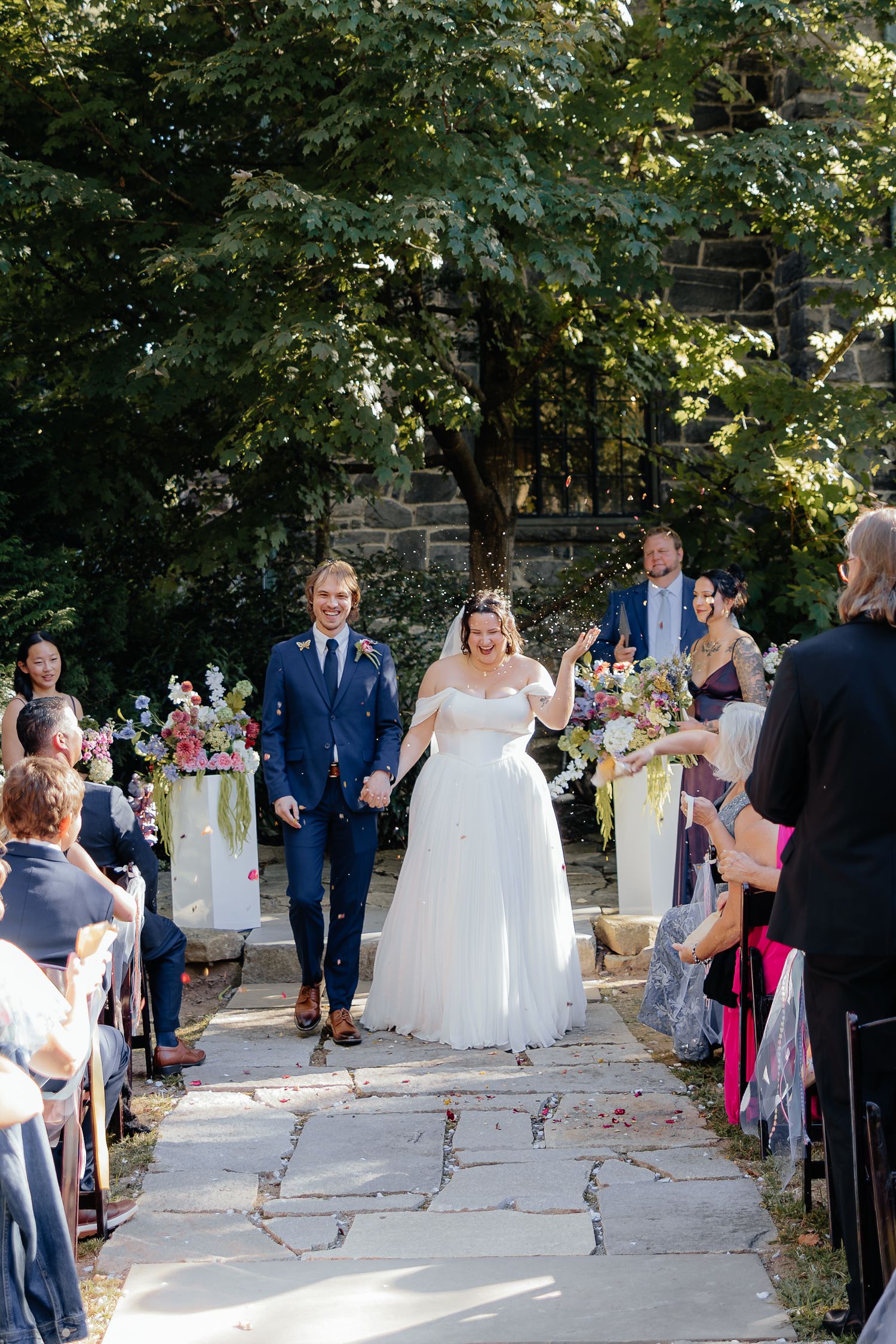 Smiling bride in a white gown and groom in a navy suit walk down a stone aisle past cheering guests at an outdoor garden wedding ceremony.