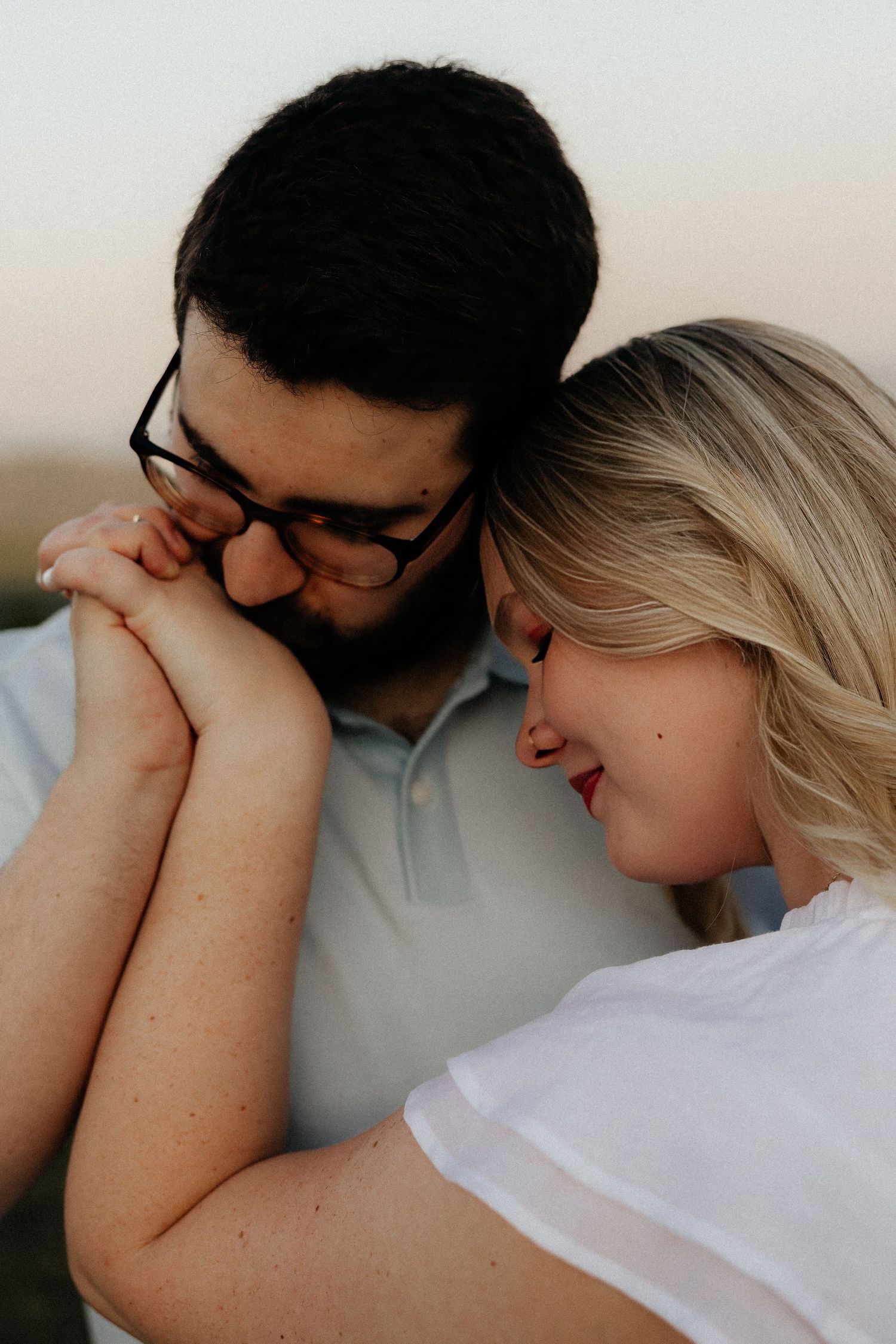 A couple shares a tender moment as a man kisses a woman's hands while she smiles softly outdoors at golden hour.