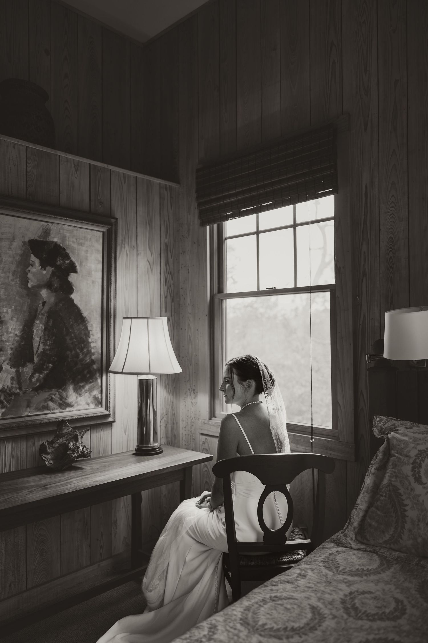 Black and white photo of a bride in white gown sitting by a window in a wood-paneled room with a portrait painting nearby.