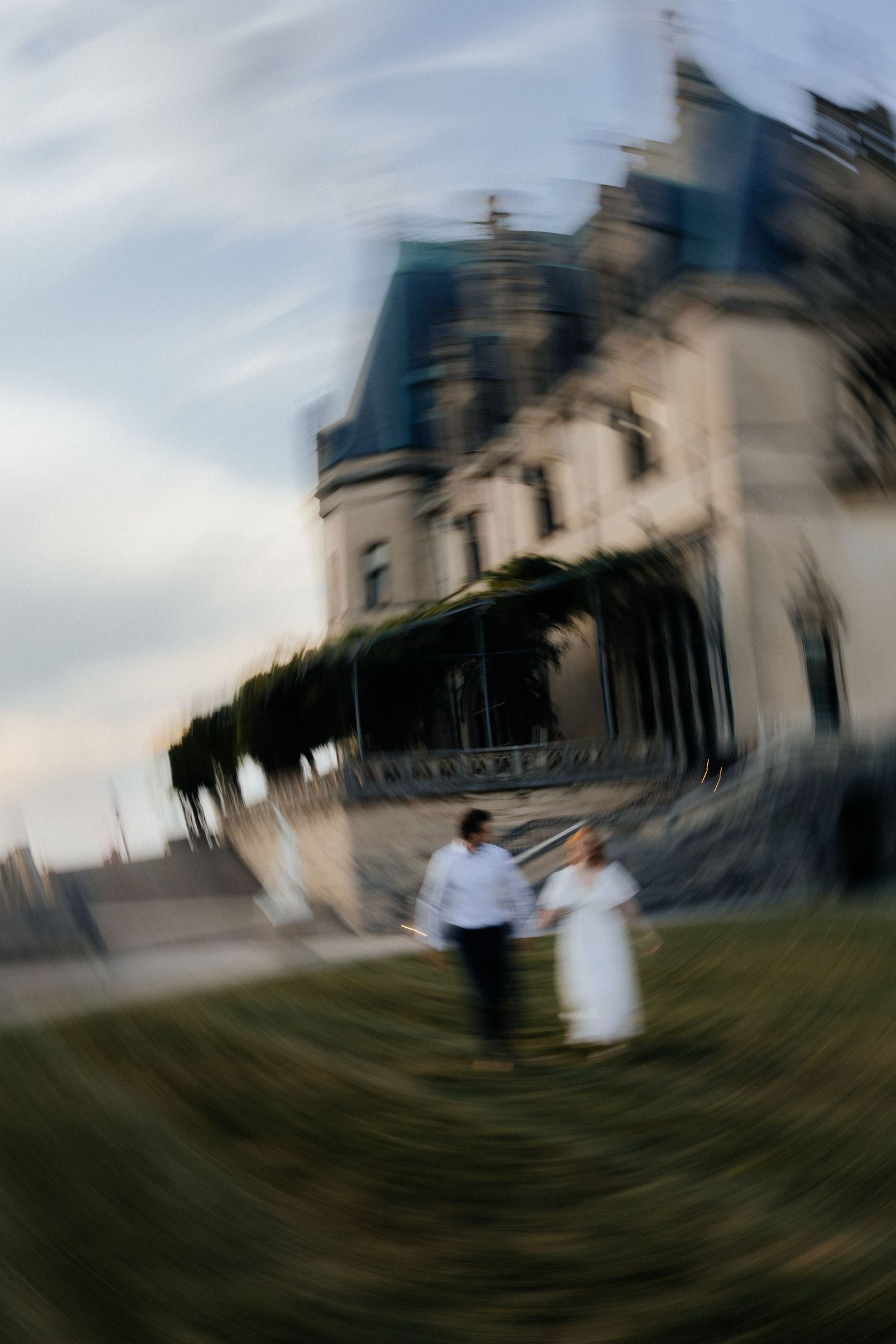 A couple in white attire holds hands while running near a grand chateau, captured with a dreamy zoom blur effect.
