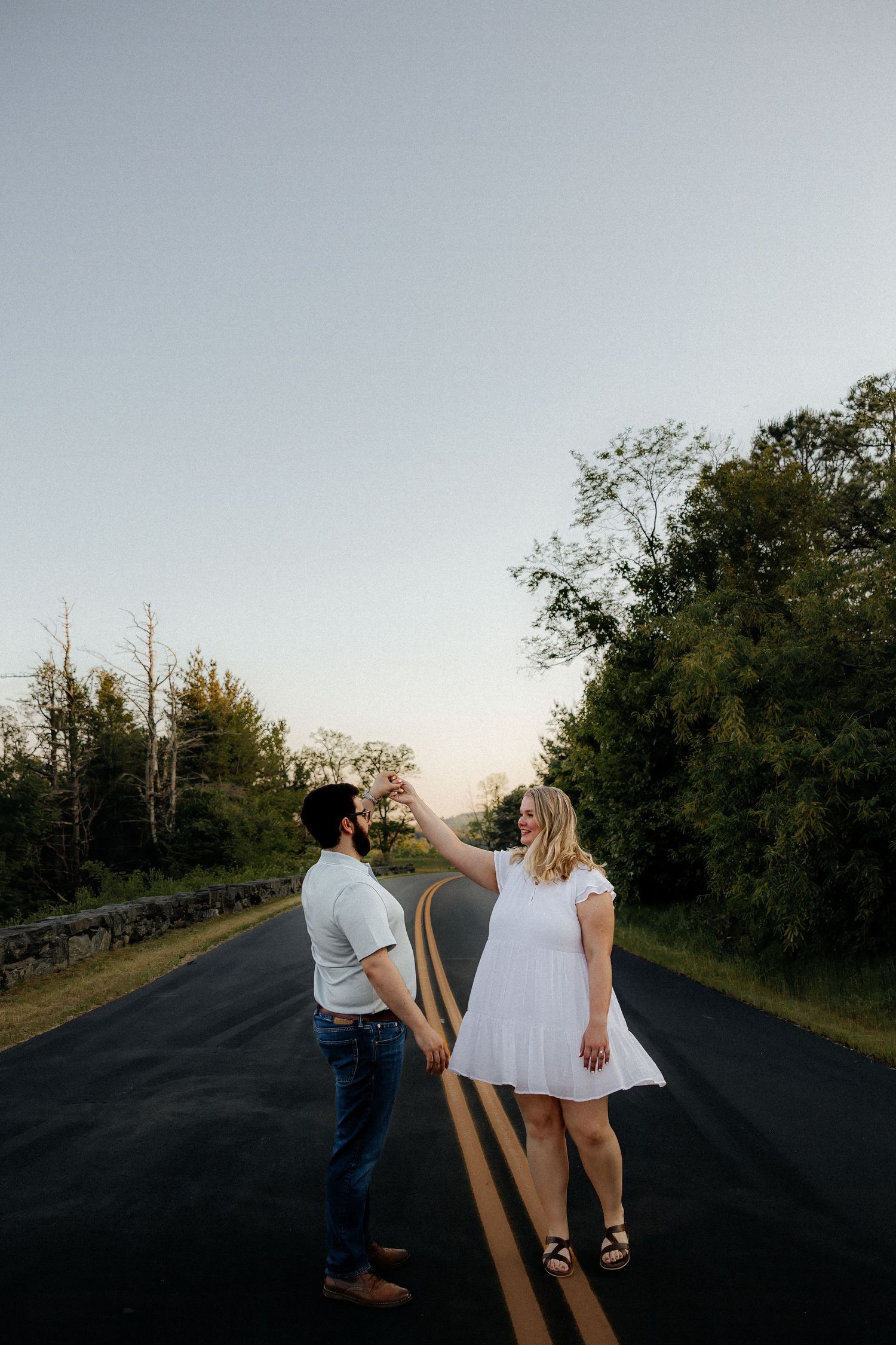 Couple dancing on empty road at sunset, woman in white dress holding man's hand among lush green trees.