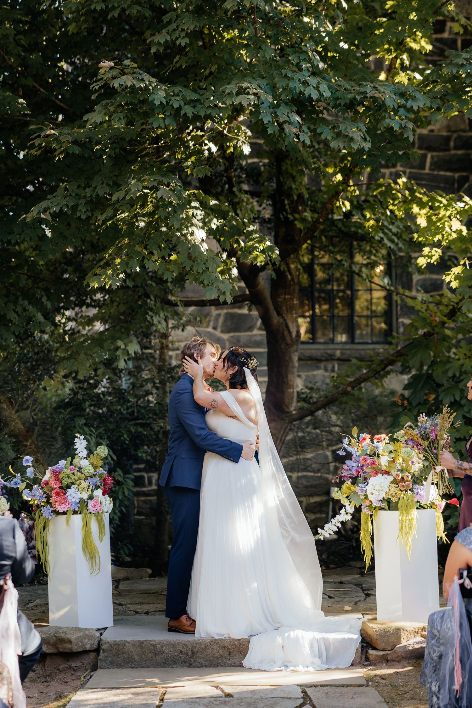 Bride and groom share first kiss at outdoor wedding ceremony surrounded by colorful floral arrangements and lush green trees.