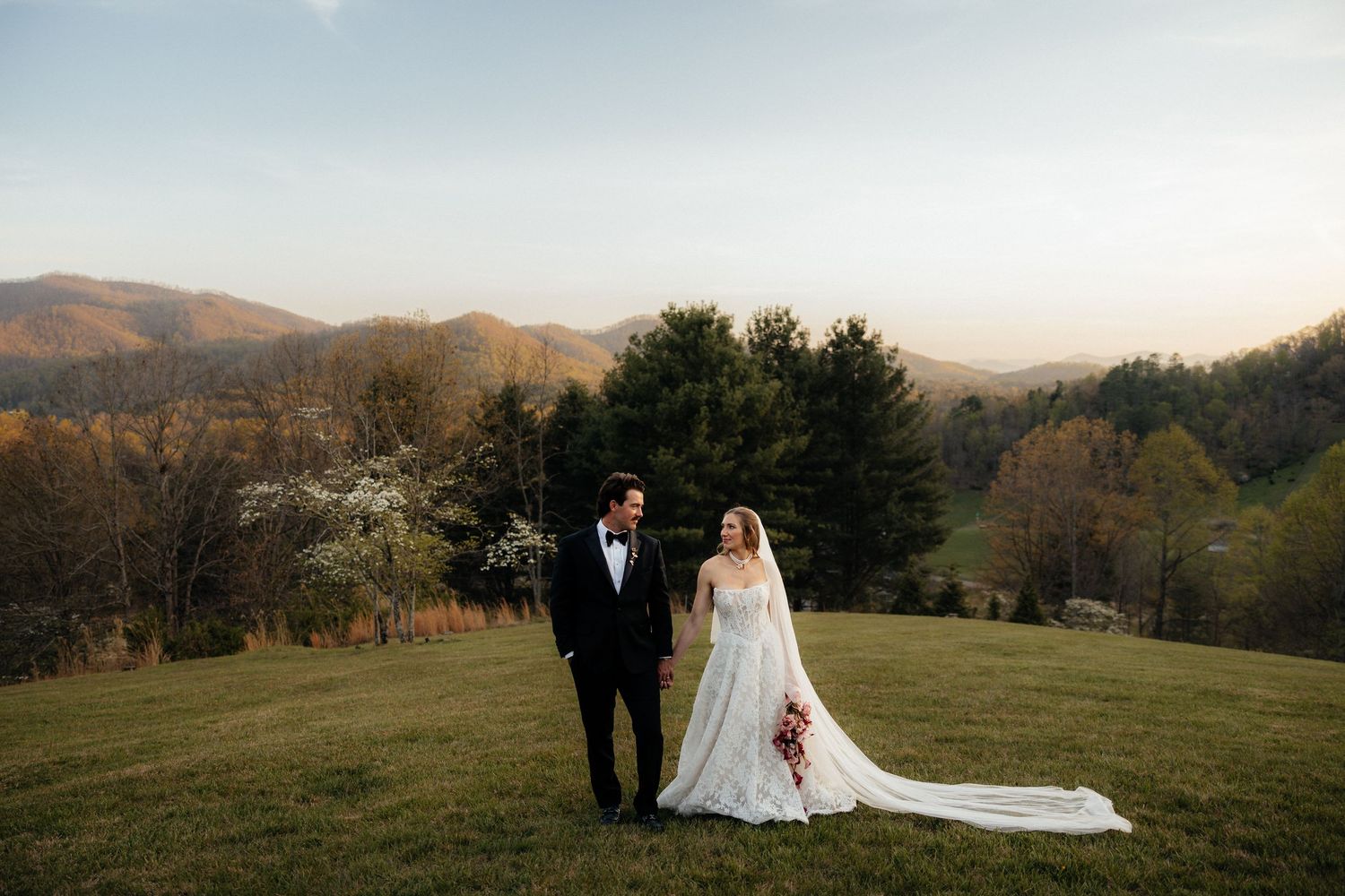 Bride and groom stand on a lush green hillside with stunning mountain views at sunset during their outdoor wedding.