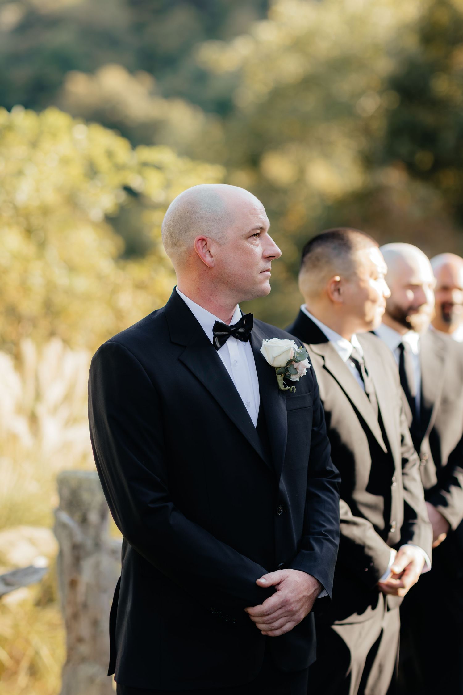 Groom in a black tuxedo with bow tie and white boutonniere stands at an outdoor wedding ceremony with groomsmen beside him.