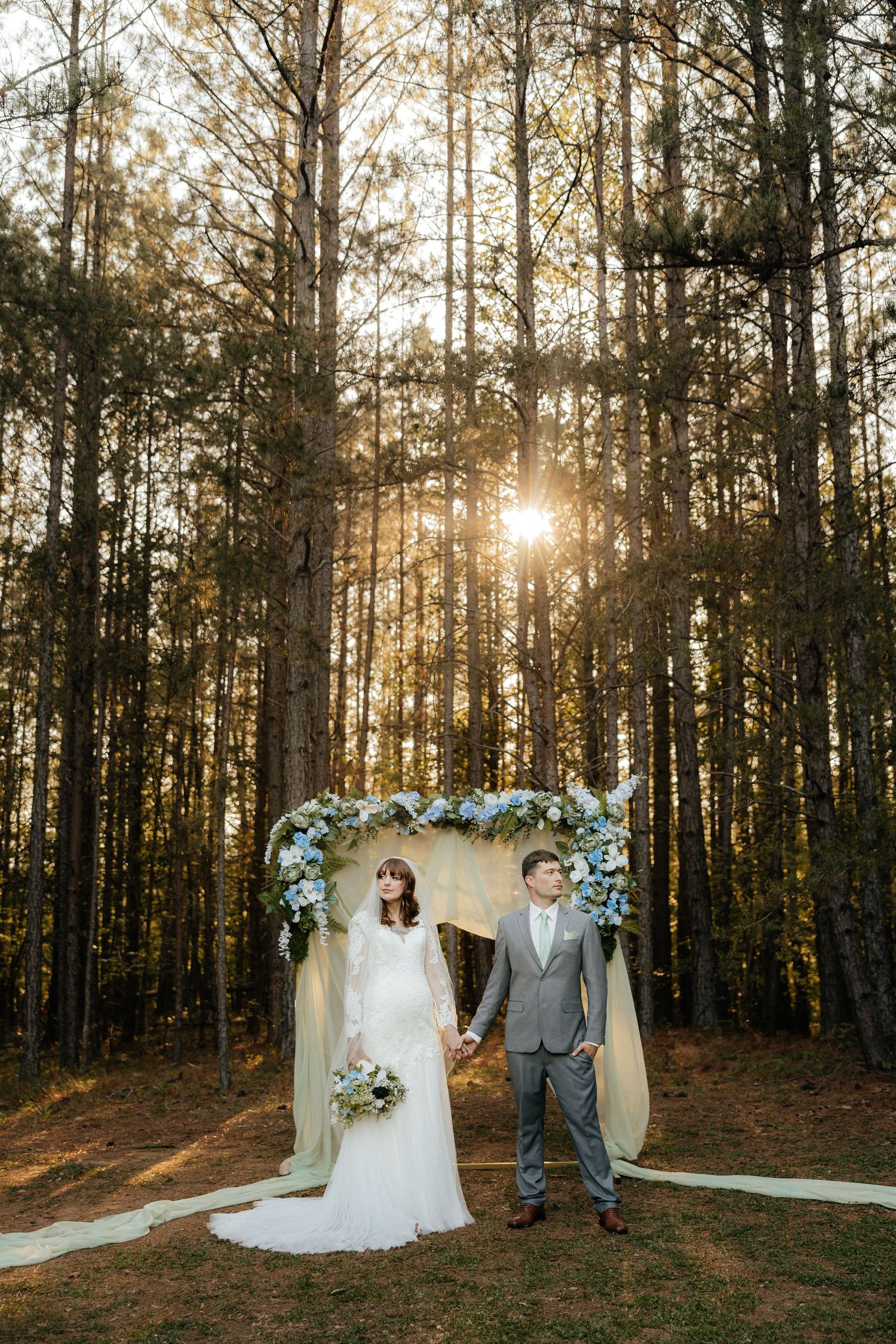 Bride and groom holding hands under a floral arch in a sunlit pine forest wedding ceremony.