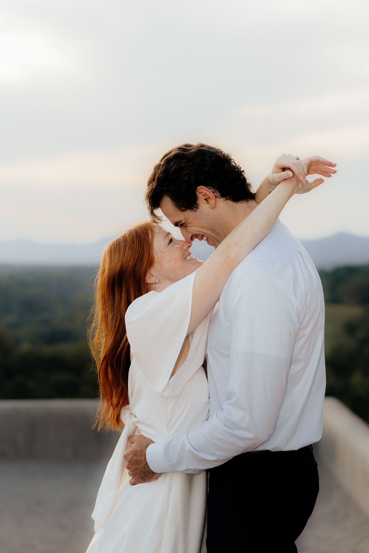 A couple in white embraces and nearly kisses on a rooftop with scenic mountain views at sunset.