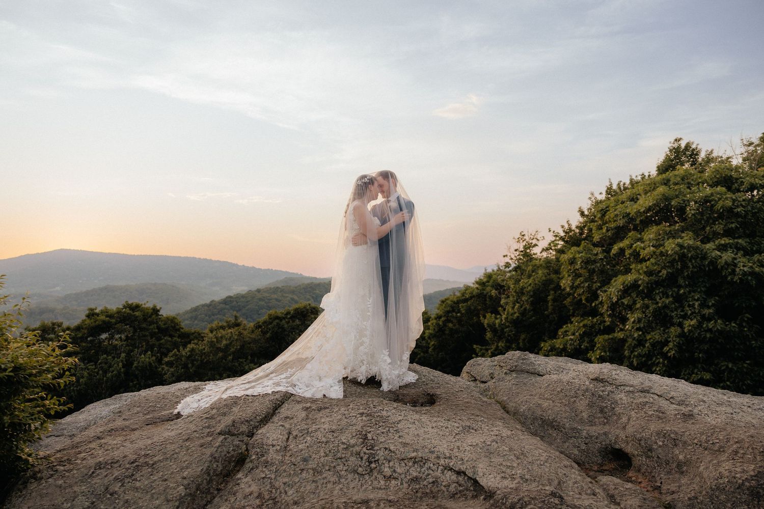 Bride and groom embrace on rocky summit at sunset with scenic mountain valley views in background.