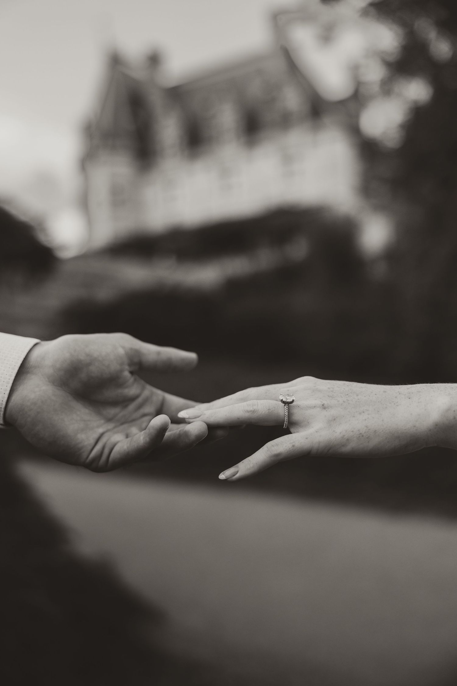 Black and white photo of two hands nearly touching, one wearing an engagement ring, with a blurred mansion in the background.